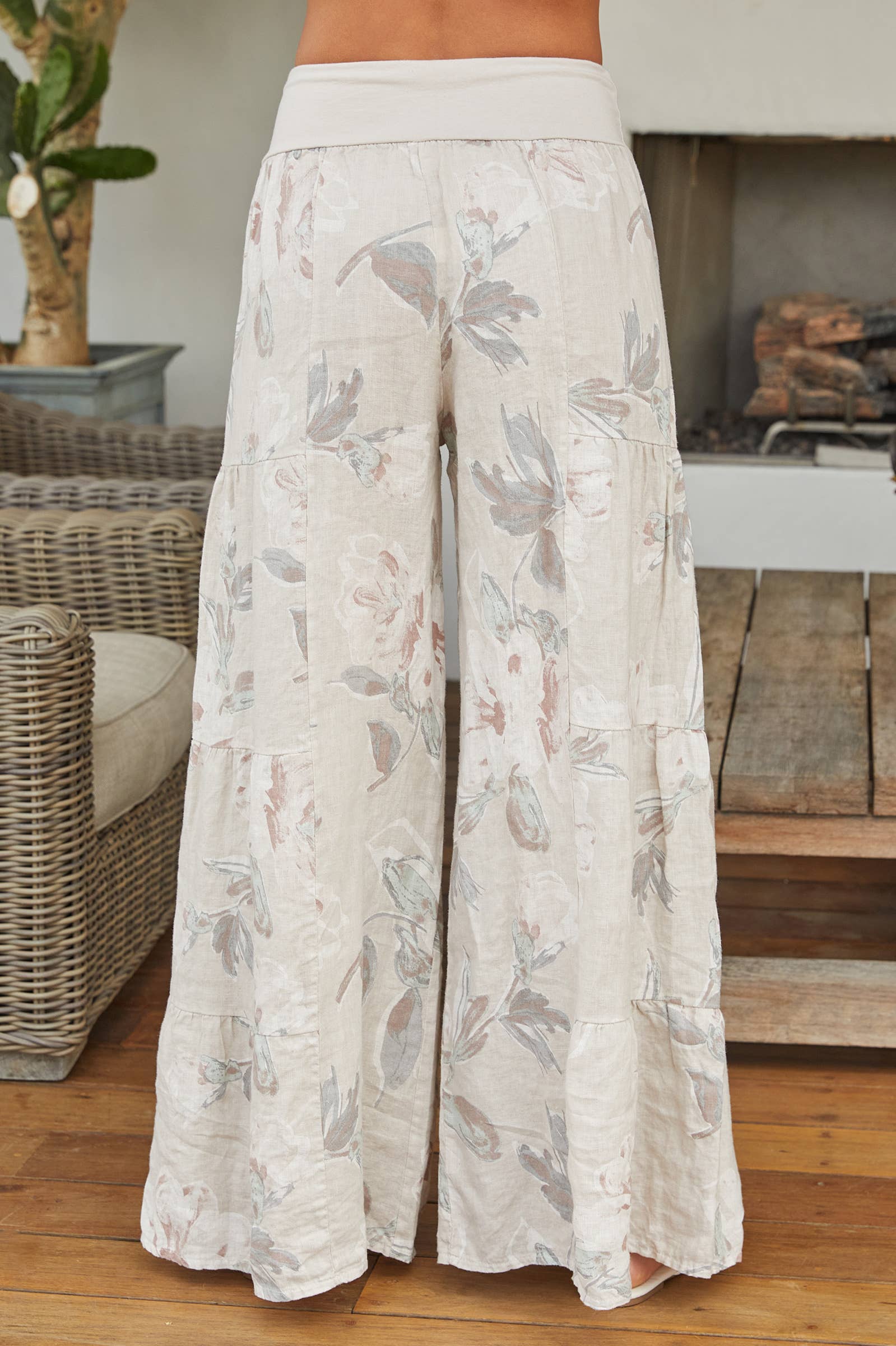 Rear view of a woman wearing cream-colored, floral-print tiered Italian linen palazzo pants with white strappy sandals.