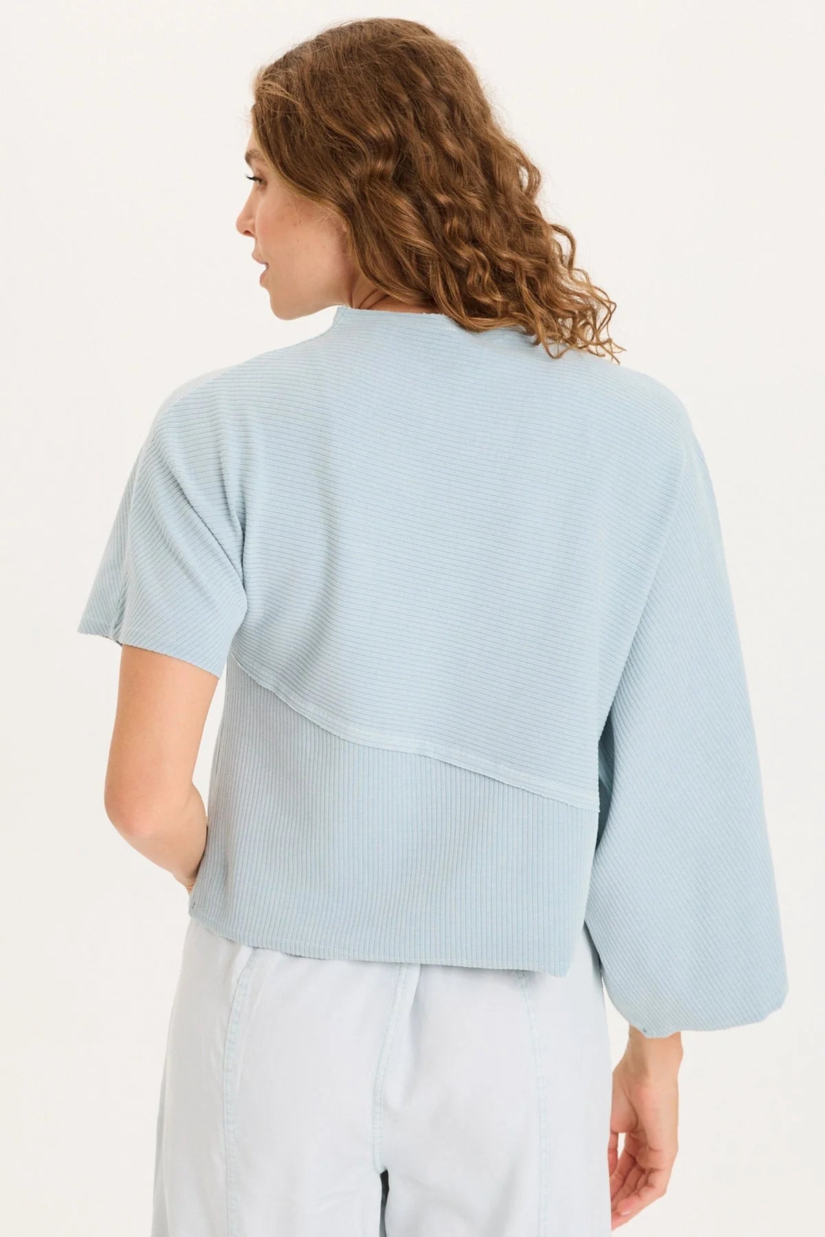 Rear view of a woman with curly hair wearing a light ice-blue asymmetrical ribbed top with one flowing sleeve and one standard short sleeve.