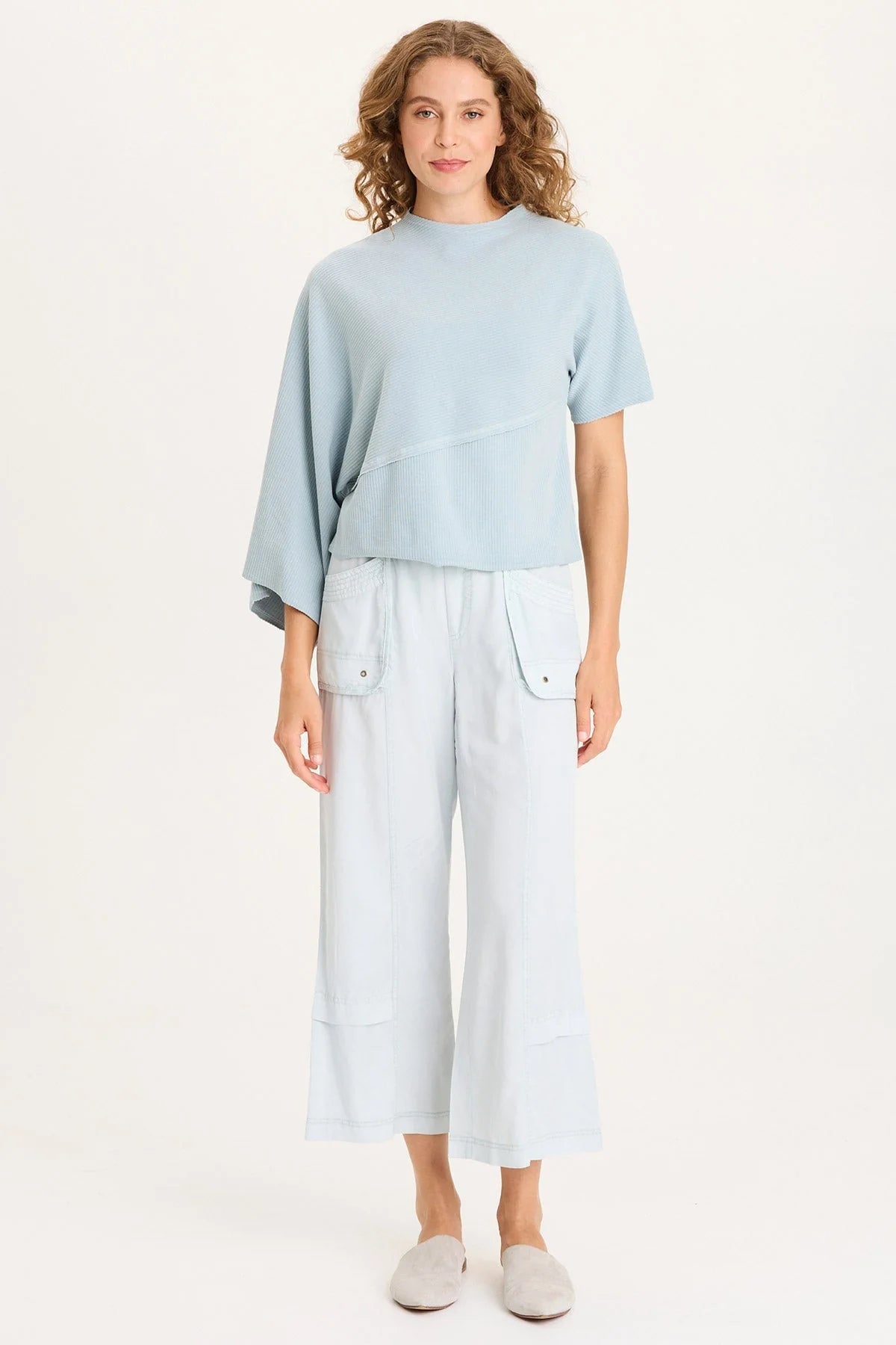 Full body of a woman with curly hair wearing a light ice-blue asymmetrical ribbed top with one flowing sleeve and one standard short sleeve.