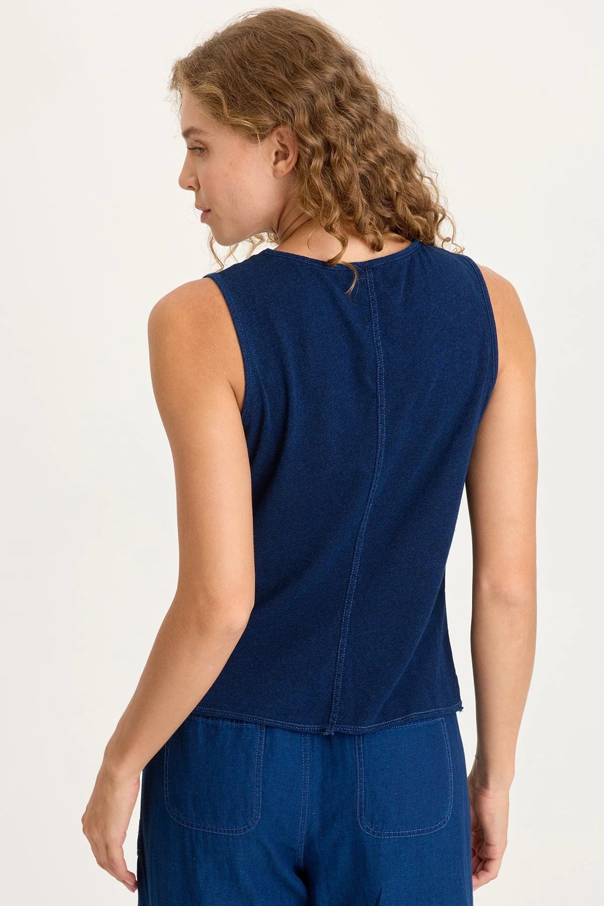 Rear view of a woman with curly blonde hair wearing a dark indigo mixed-media tank top featuring adjustable front cinch cords and a raw-edge hem.