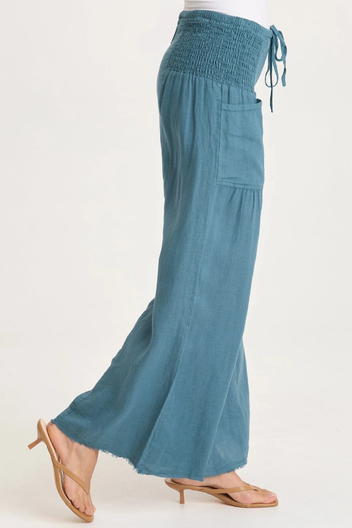 Side view of a woman wearing flowing teal-blue wide-leg linen pants with unique smocked side waist panels and deep front pockets.