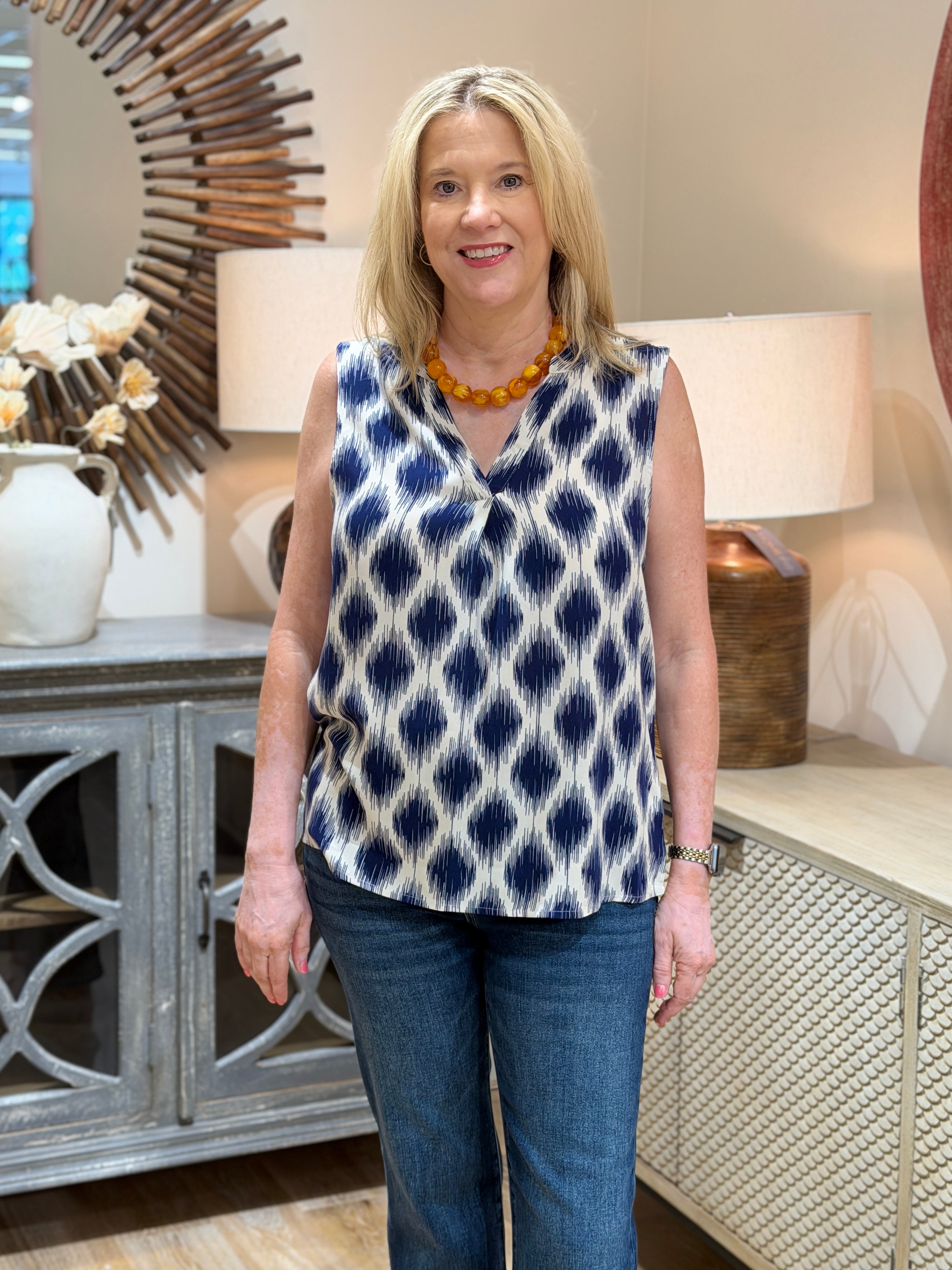 Blonde woman wearing a navy and cream geometric ikat print sleeveless blouse with blue jeans.