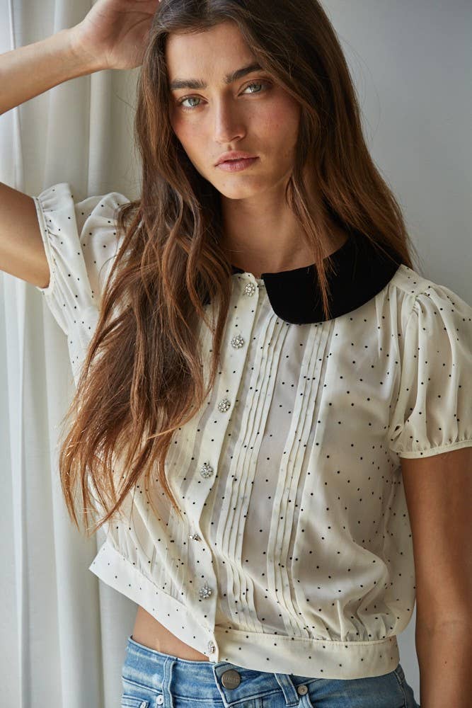 Woman wearing the black and white polka dot blouse with a velvet Peter Pan collar and a blue jean standing near a window.