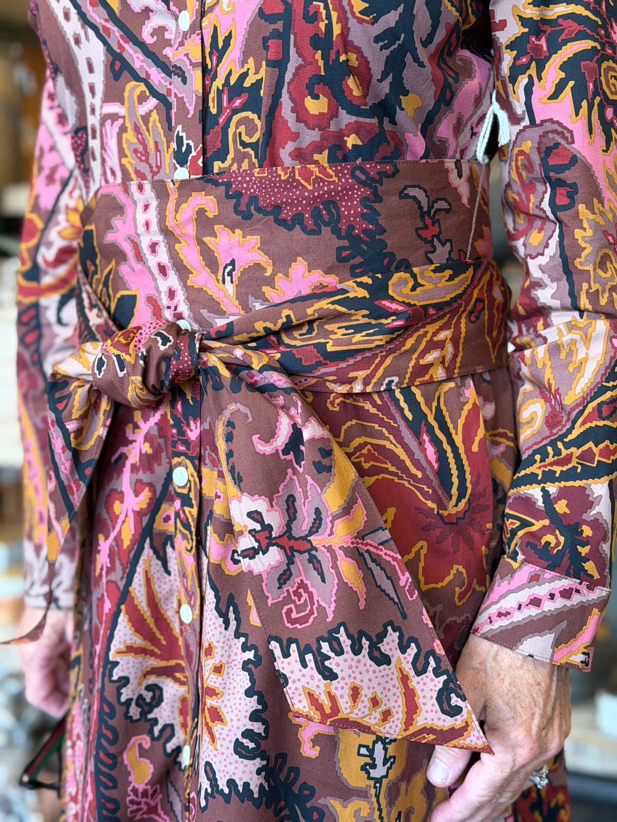 A close-up of a person wearing a long-sleeve belted wrap dress with a vibrant, multicolored paisley and floral pattern in pink, brown, orange, and yellow.