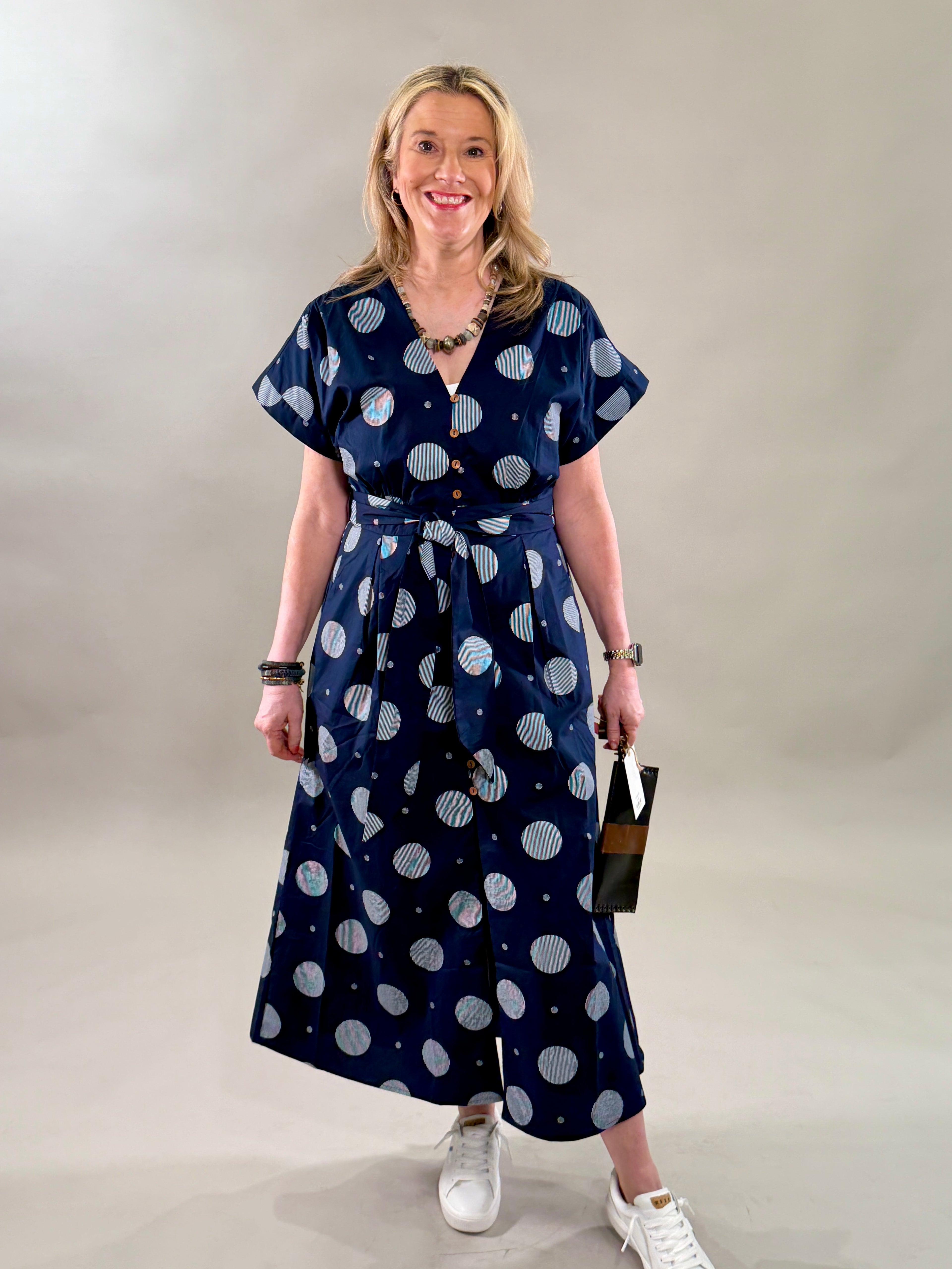 Woman smiling in a navy polka dot 100% cotton midi dress with a removable belt and white sneakers.