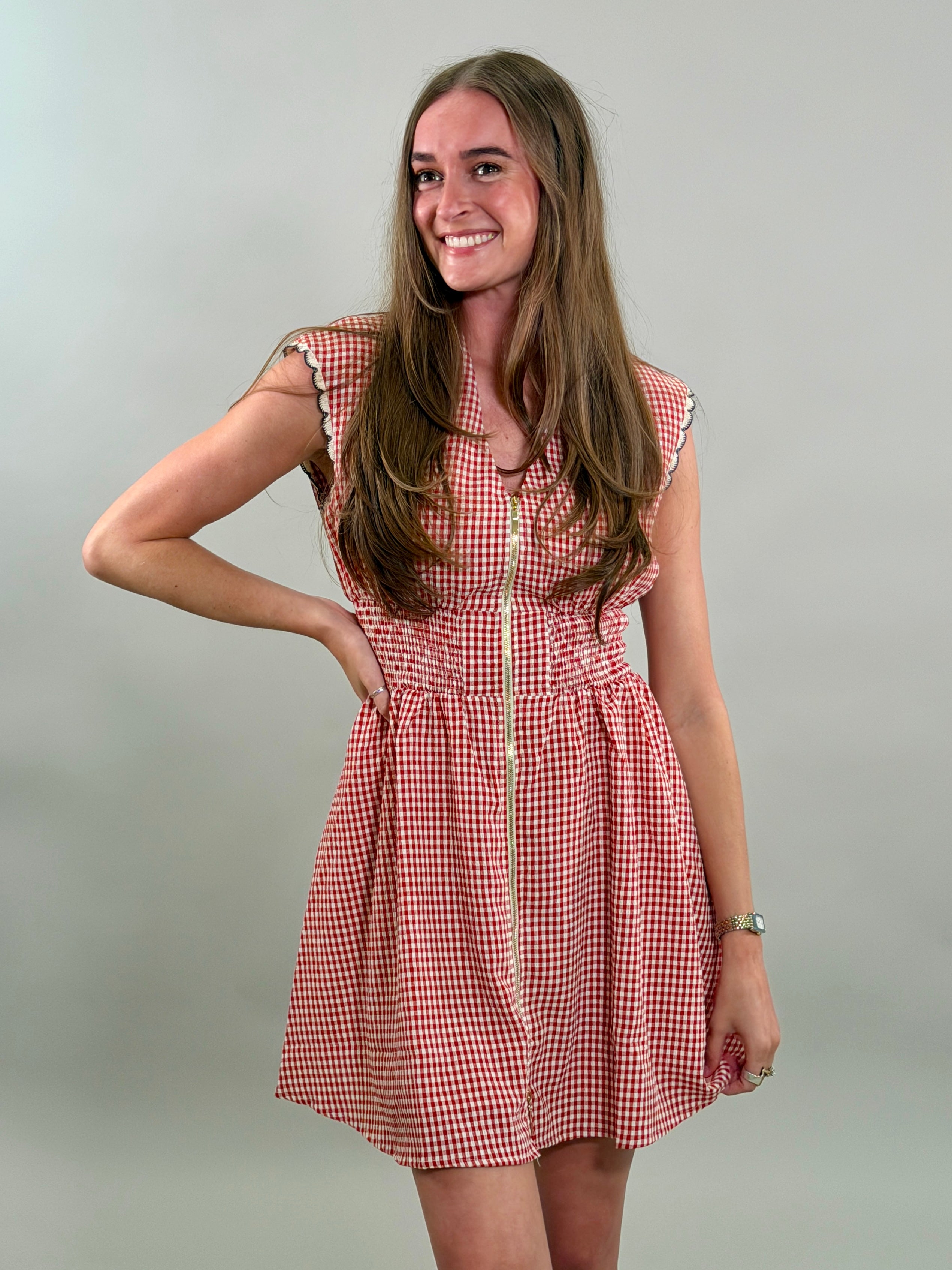 A smiling woman looking to the side with long brown hair wearing a red and white gingham sleeveless dress with a gold front zipper and a smocked waistband.
