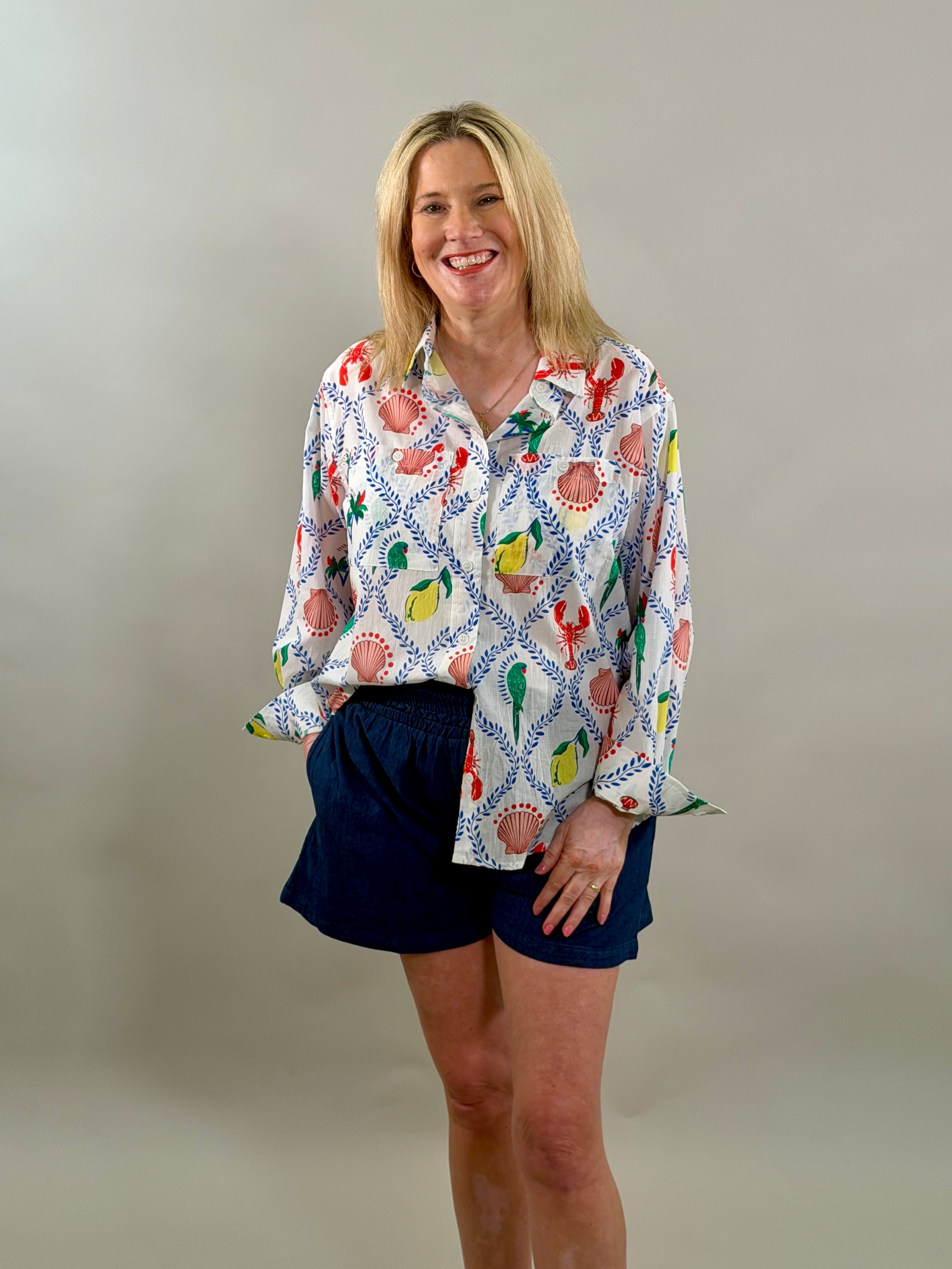 Smiling woman wearing an oversized white button-down shirt with a vibrant blue, red, and yellow coastal print paired with navy shorts.