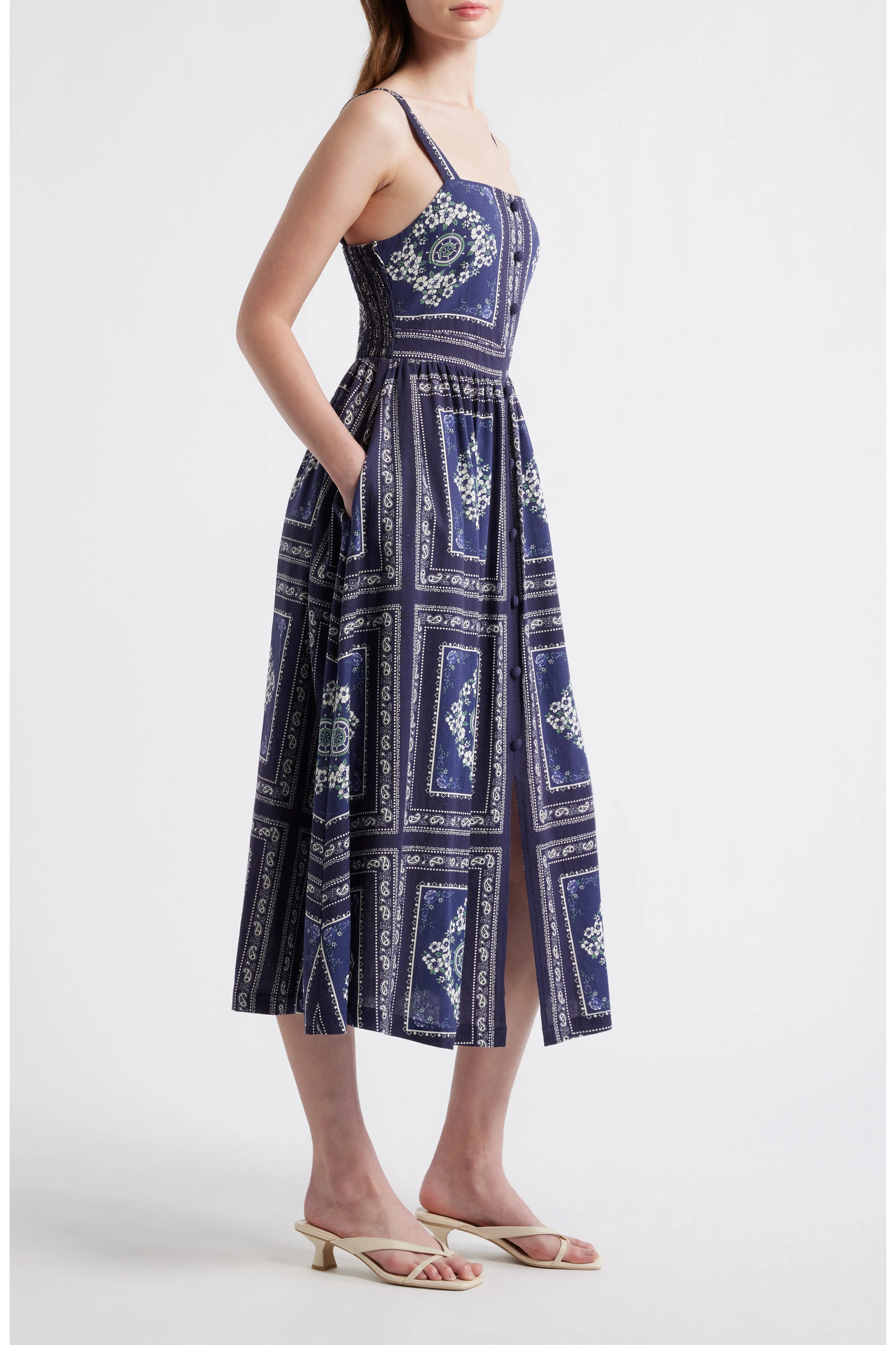Side view of a woman wearing a navy blue bandana-patterned cotton-linen midi dress with a square neckline, adjustable straps, and a button-front skirt.