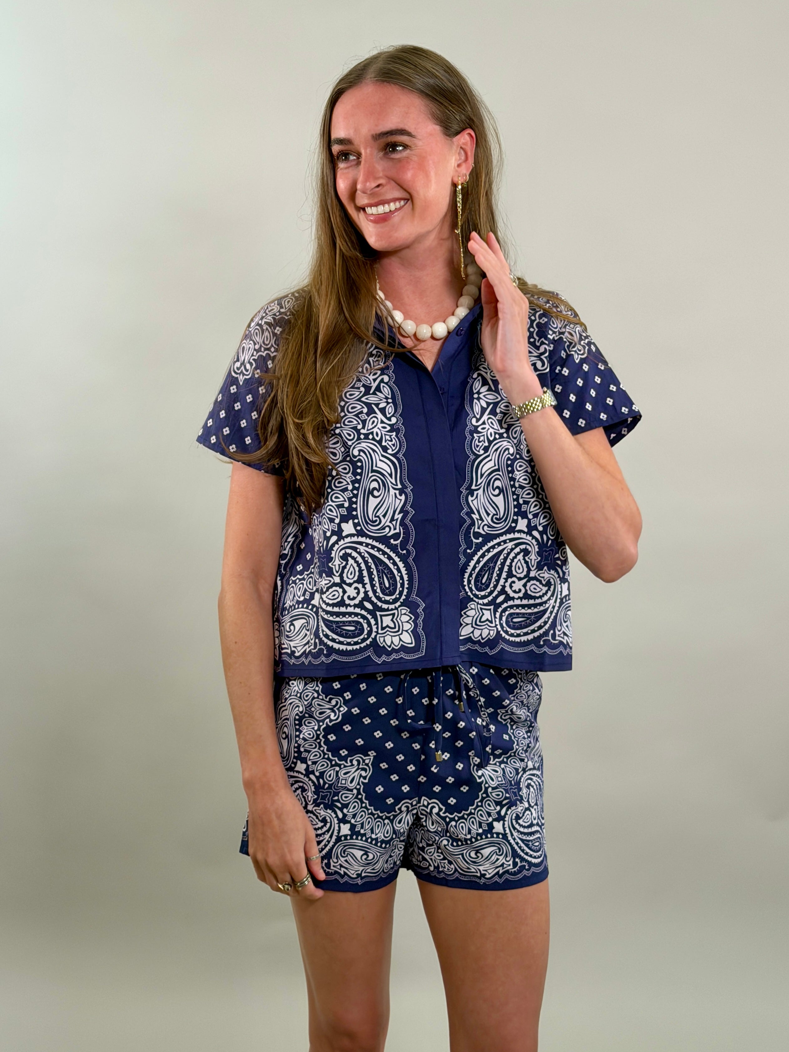 A smiling woman with long brown hair looks to her side while wearing a navy blue and white paisley bandana-print short-sleeve button-down shirt paired with matching printed drawstring shorts.