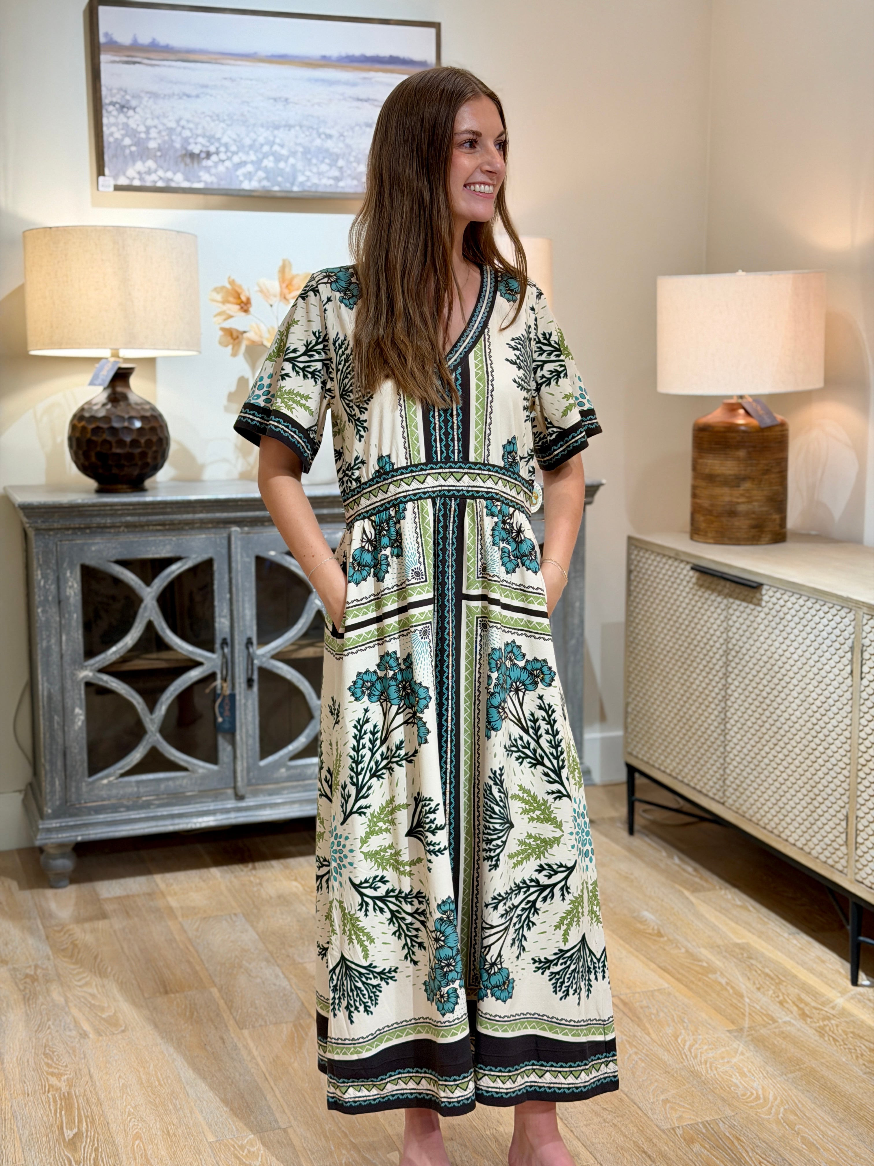 A woman modeling a short-sleeve maxi dress with a botanical print in teal, green, and brown on an off-white background, standing in a styled room with wood floors and sideboards.