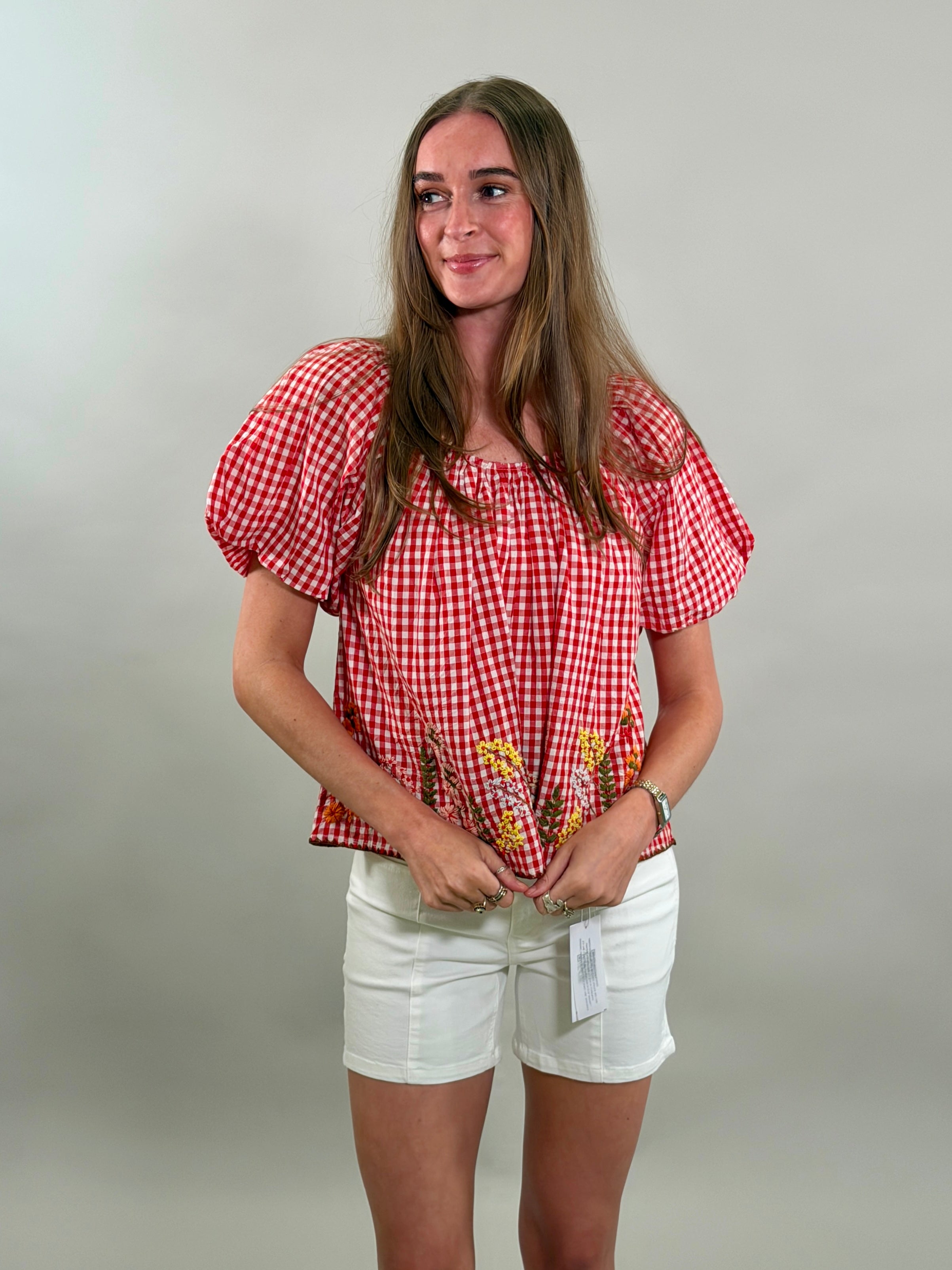 Woman looking to the side wearing a red checkered blouse with puff sleeves and white shorts against a plain background