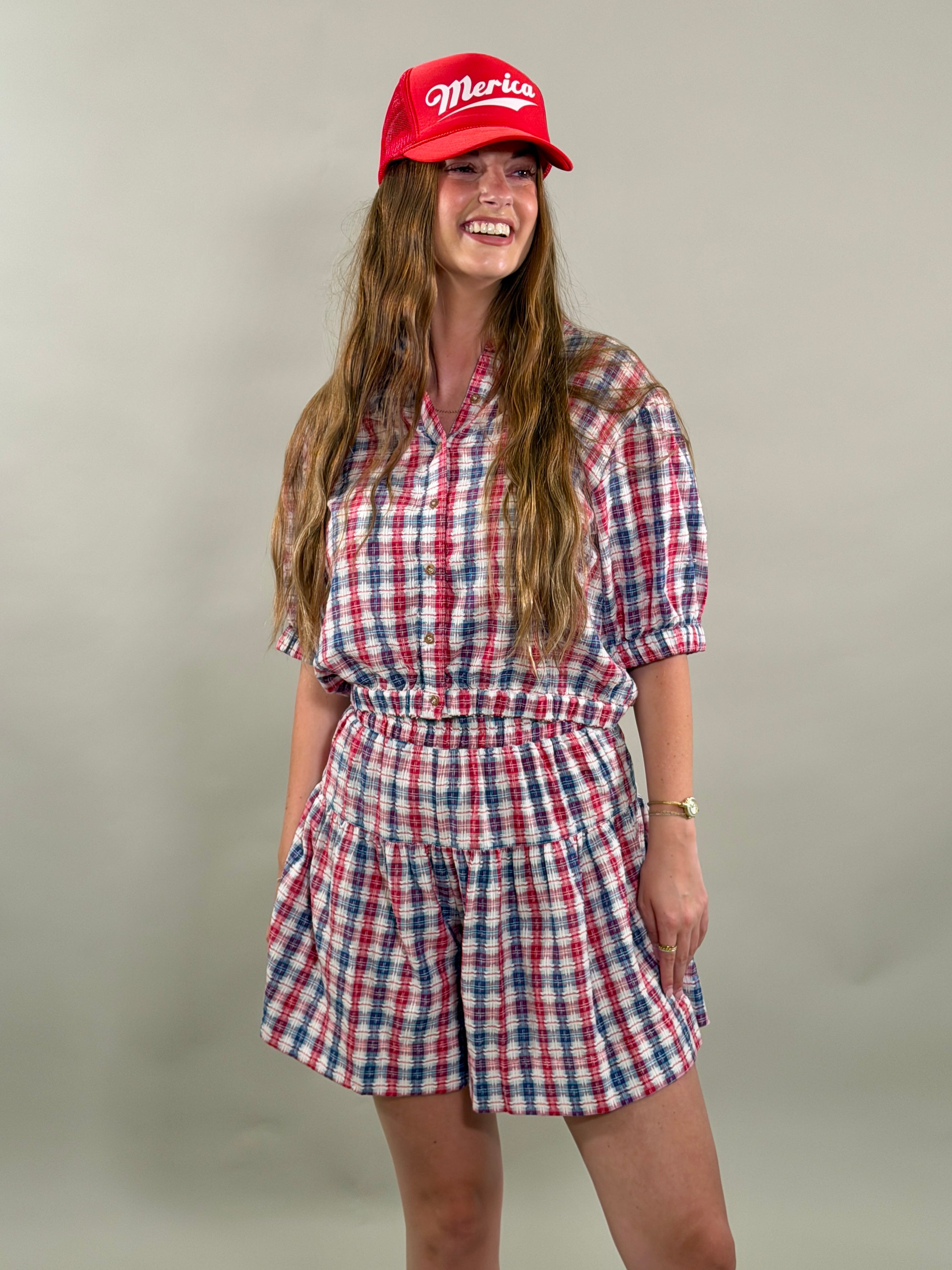 Woman smiling and looking to the side, wearing a red, navy, and white plaid short-sleeve cropped button-down shirt with a matching skort and a red baseball cap.