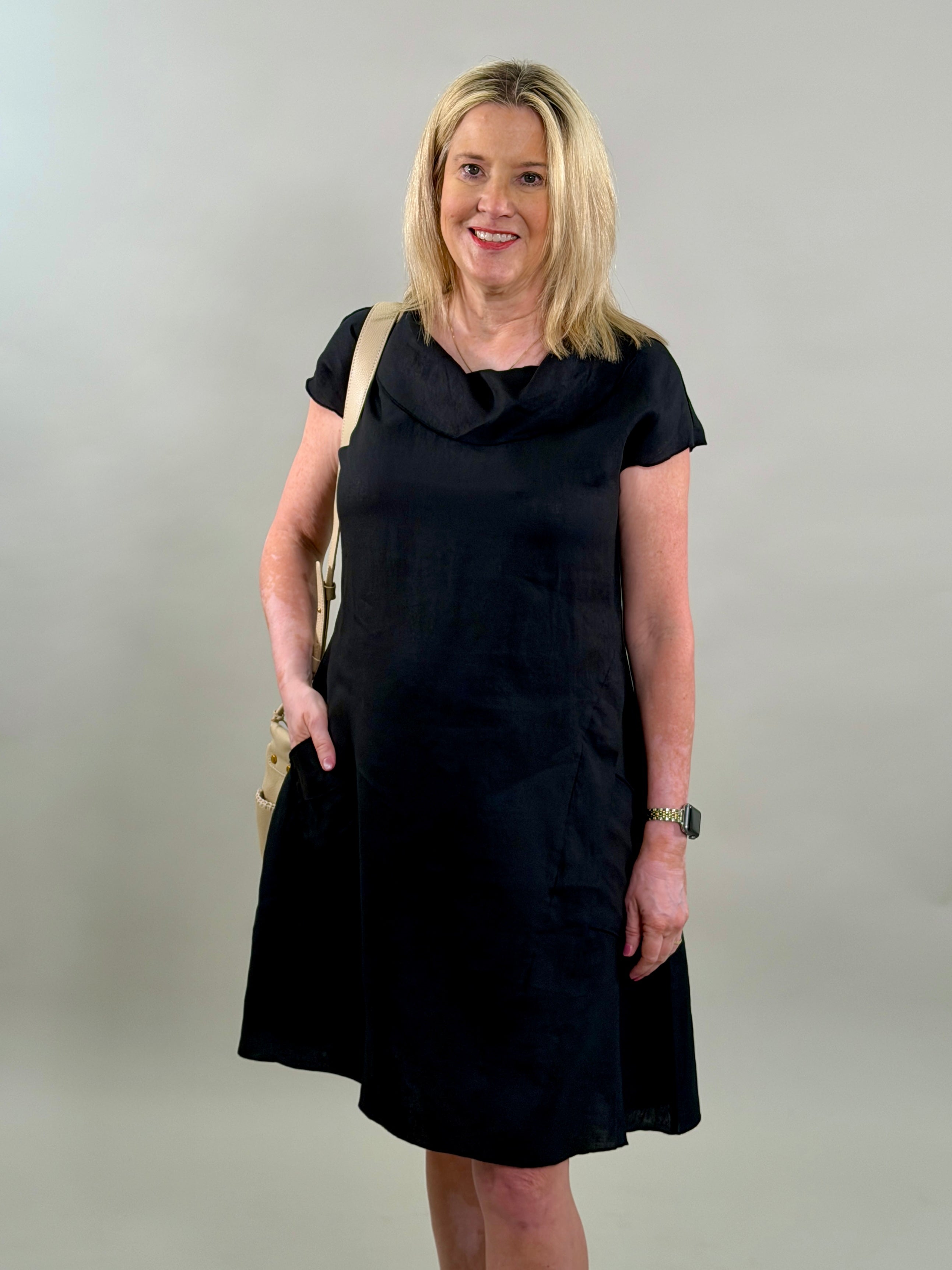 Blonde woman smiling while wearing a classic black 100% linen cowl-neck midi dress with a tan crossbody bag and hand in her pocket.