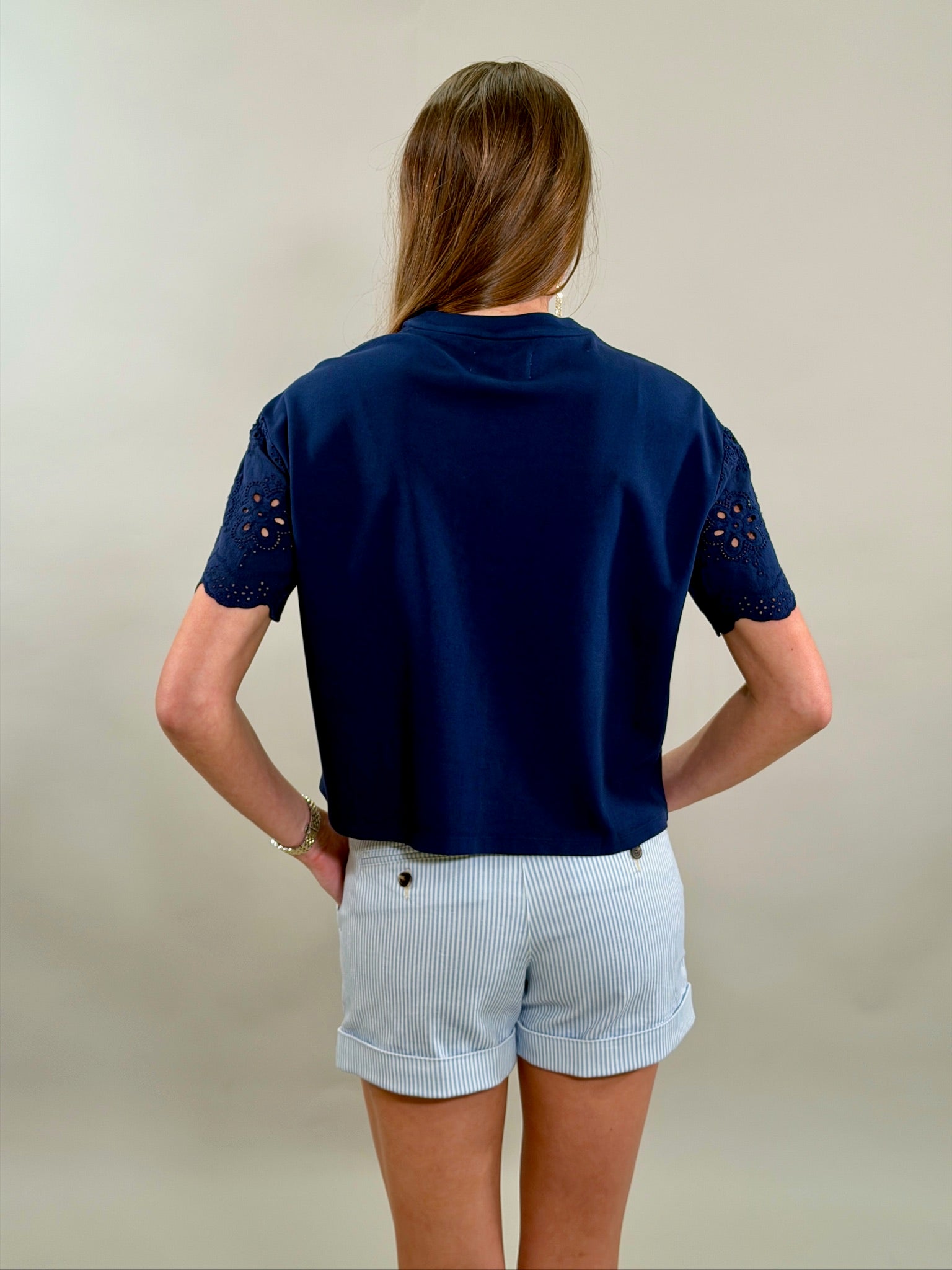 Rear view of a woman wearing a navy blue top with eyelet sleeves and light blue pinstripe shorts on a plain background