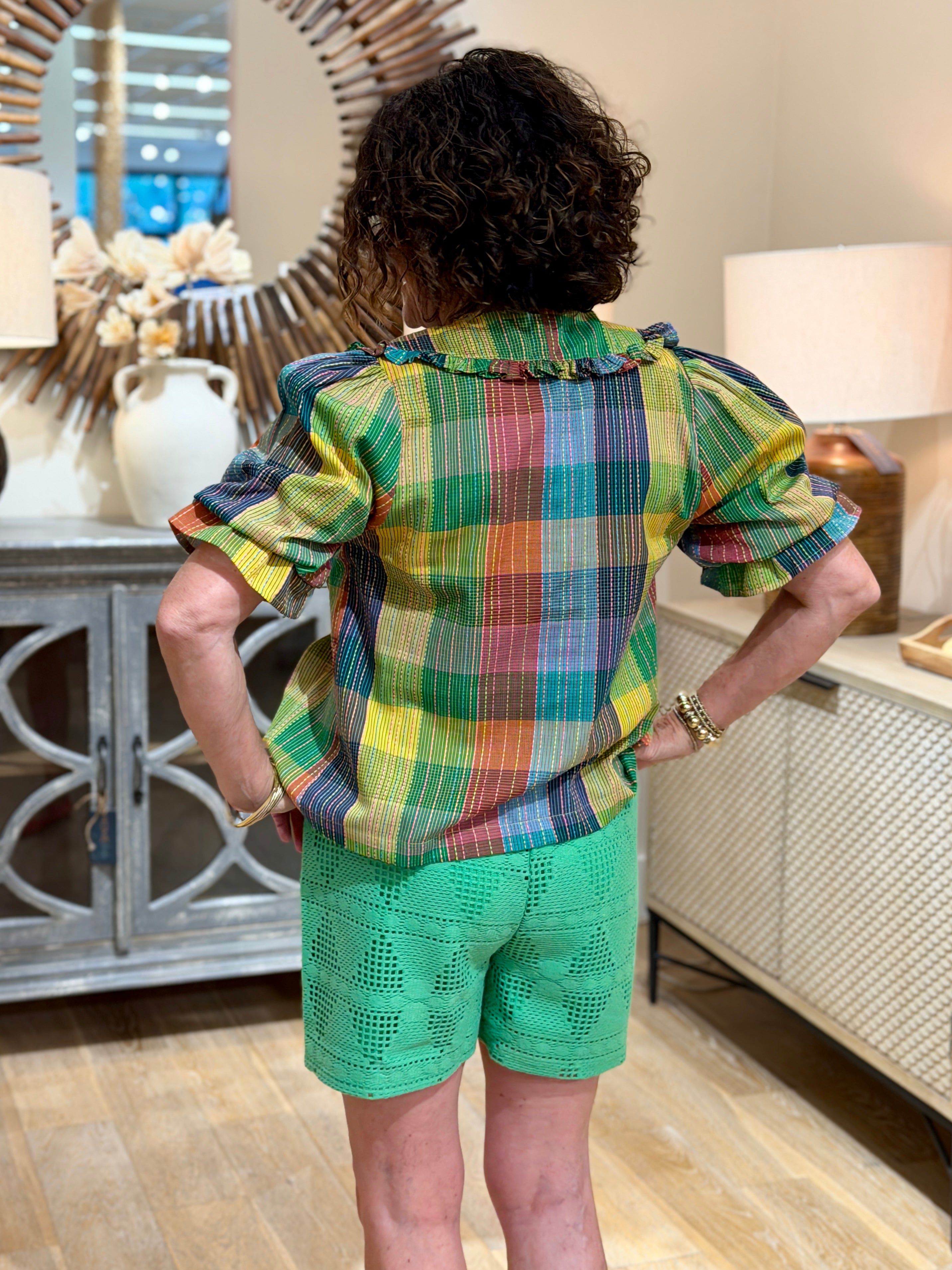 Close up of the rear of a woman with curly hair wearing a multi-colored plaid short sleeve blouse with a large ruffled collar and green eyelet shorts.