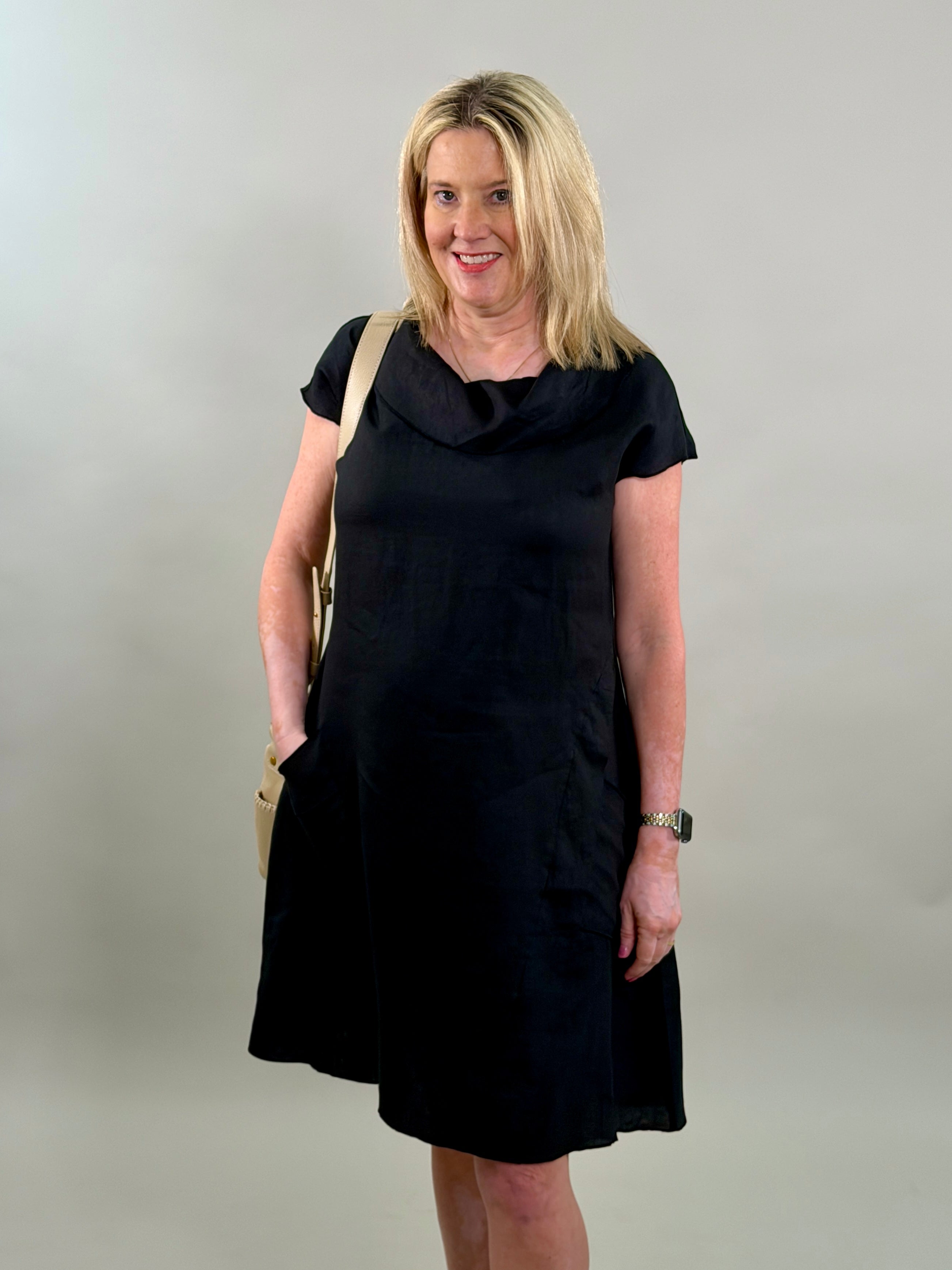 Blonde woman smiling while wearing a classic black 100% linen cowl-neck midi dress with a tan crossbody bag.