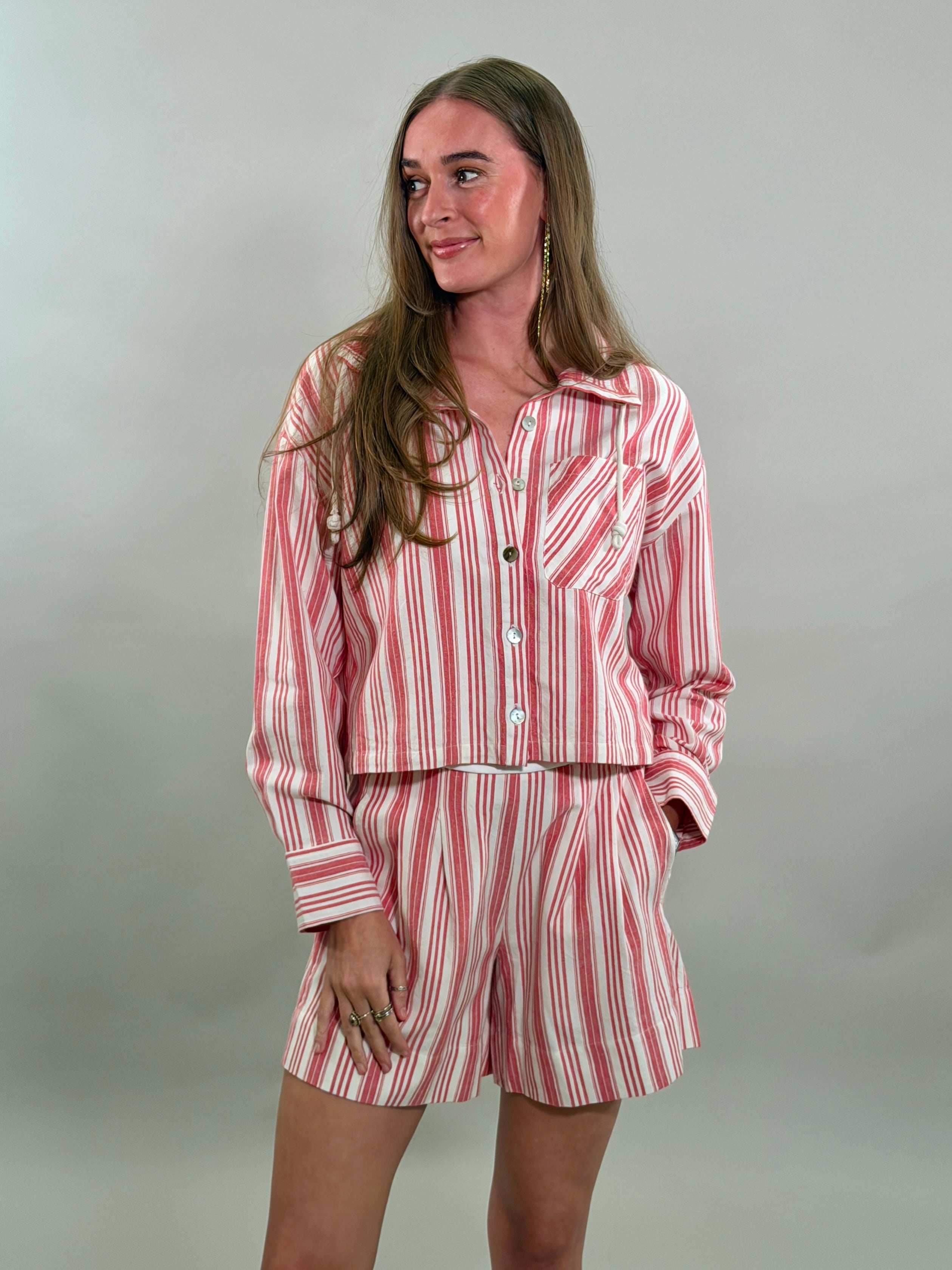 A smiling woman with long brown hair wearing a matching red and white candy-striped cropped long-sleeve shirt and pleated shorts.