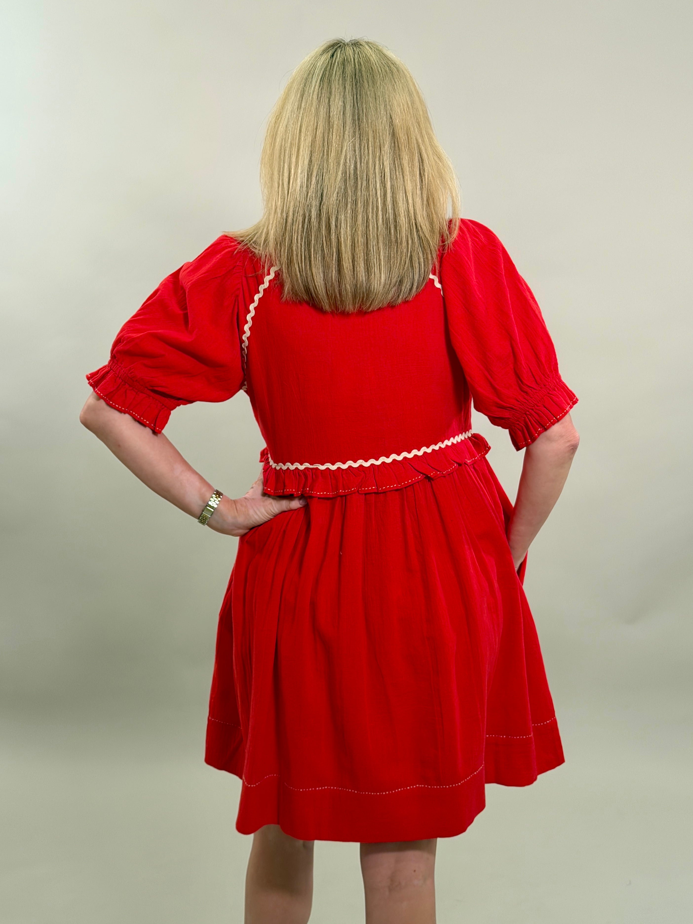 Rear view of a blonde woman with her hand on her hip wearing a vibrant red 100% cotton dress with cream-colored ricrac embroidery and puff sleeves.