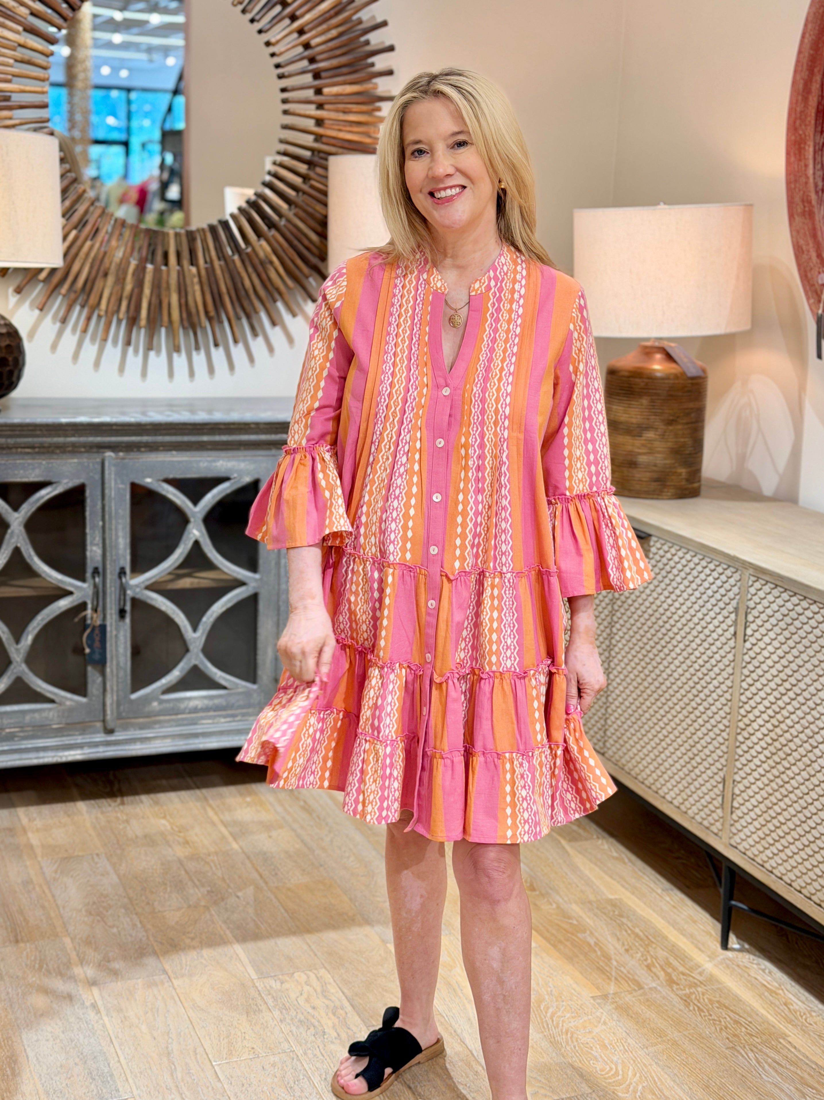 A blonde woman smiling while wearing a pink and orange patterned 100% cotton tiered dress with ruffle sleeves and black slides.