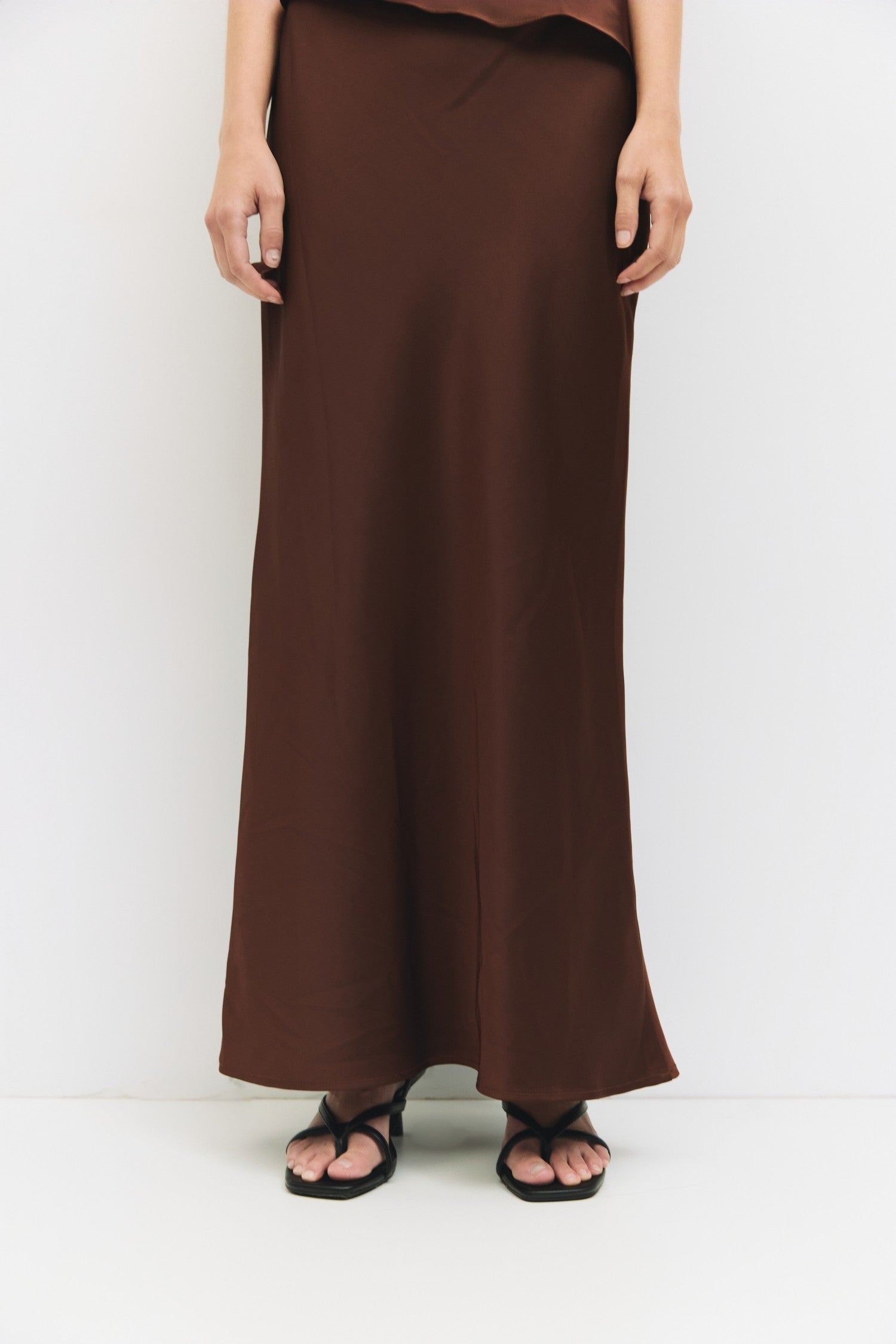 Close up of a woman wearing a chocolate brown bias-cut satin midi skirt paired with black strappy sandals.