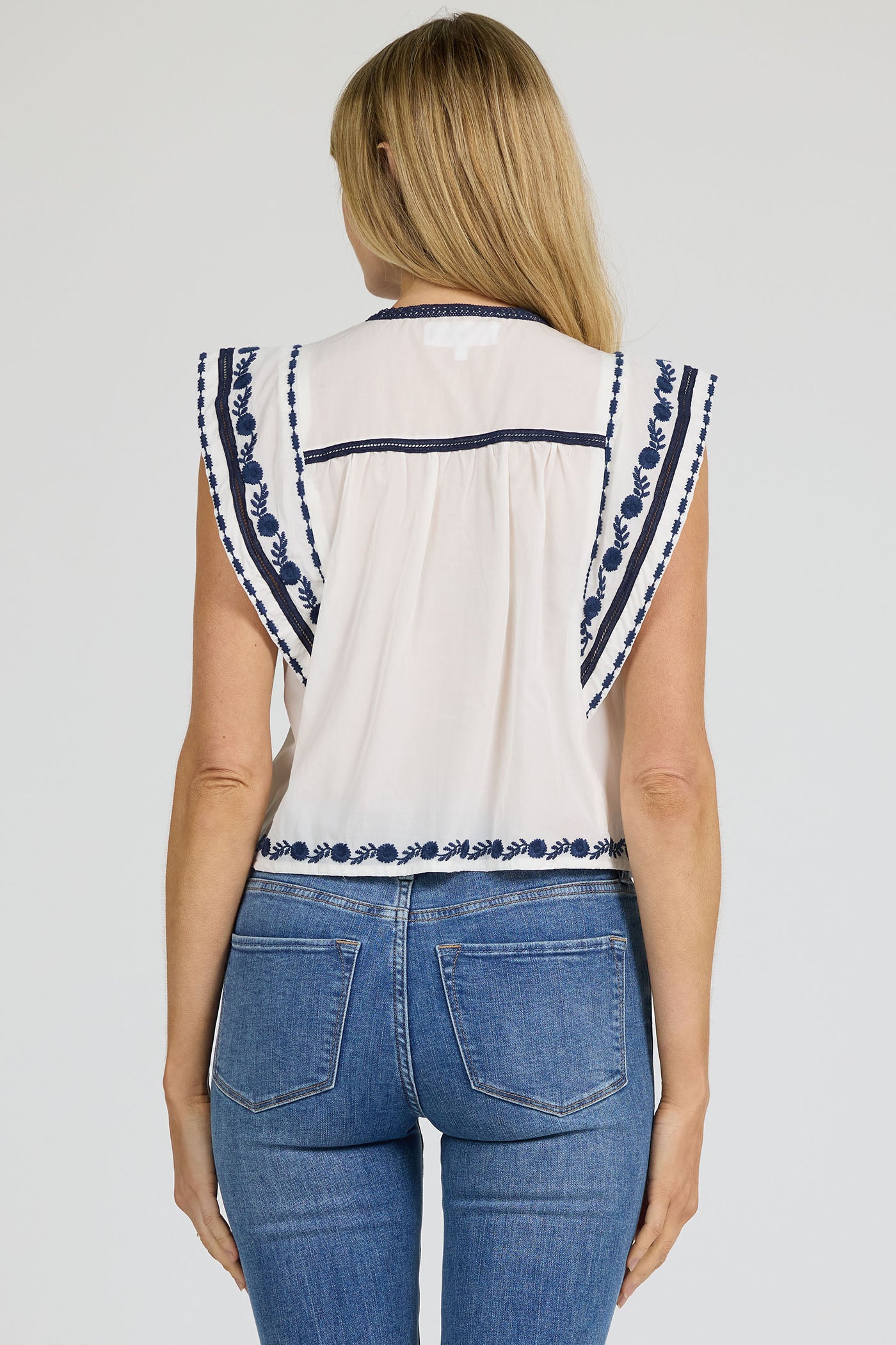 Rear view of a woman wearing a white sleeveless split-neck top with blue embroidery and blue jeans