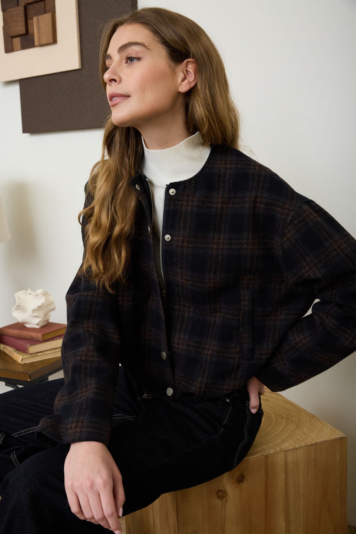 A woman sitting wearing a navy and brown plaid collarless bomber jacket layered over a white turtleneck sweater with dark jeans.