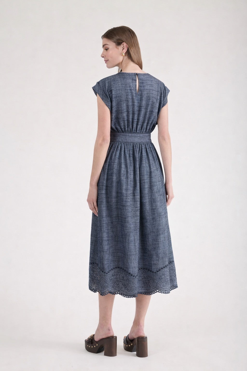 Rear view of a woman wearing a short-sleeve chambray midi dress with a wide grommet belt, a gathered waist, and an intricate floral eyelet scalloped hem.