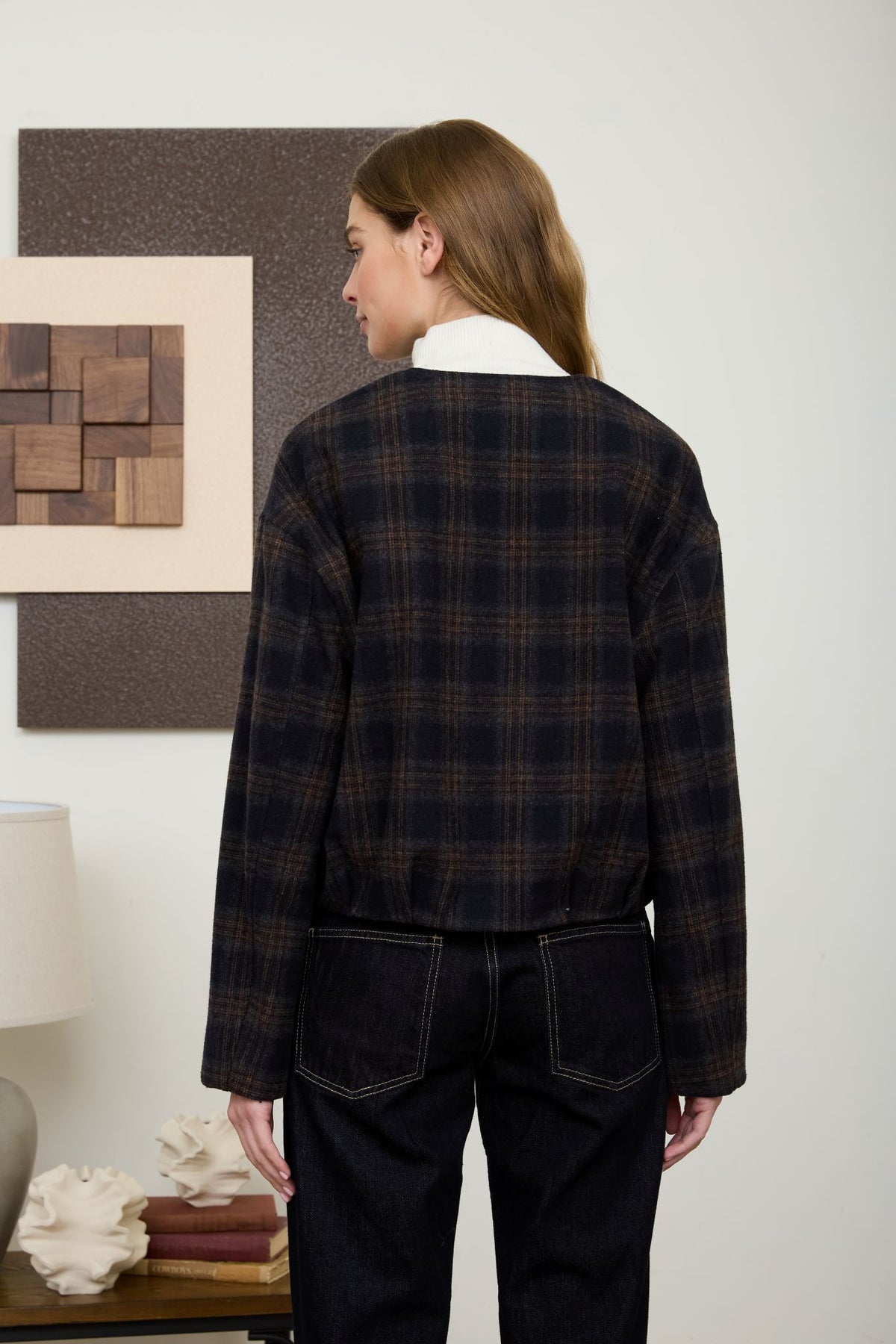Rear view of a woman wearing a navy and brown plaid collarless bomber jacket layered over a white turtleneck sweater with dark jeans.