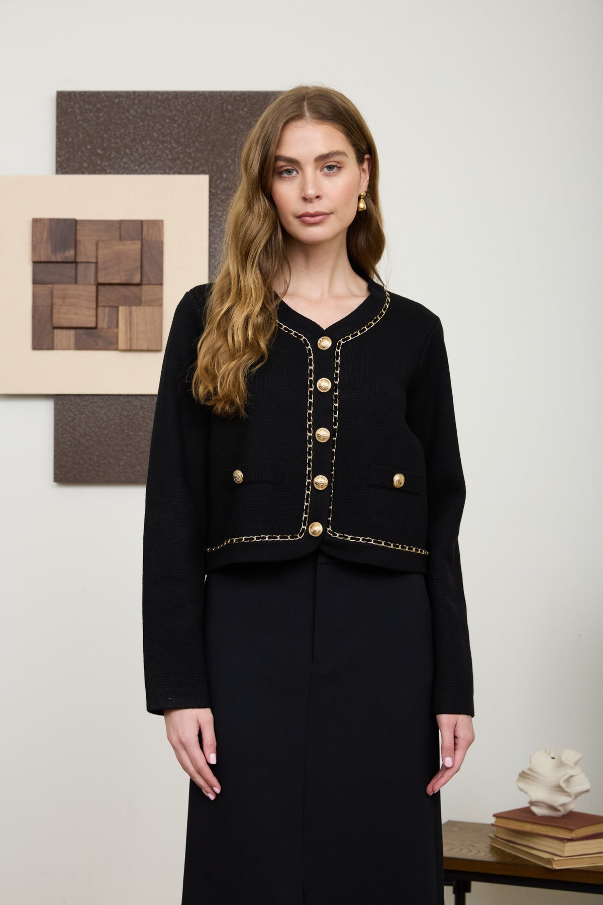 A woman wearing a structured, long-sleeve black knit cardigan featuring a V-neckline, prominent gold crest buttons, and intricate gold chain-link trim.