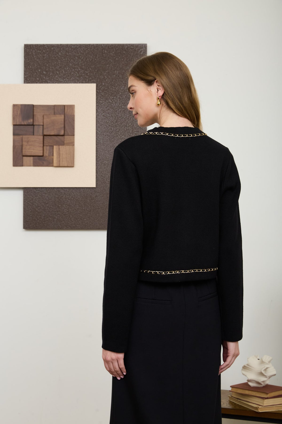Rear view of a woman wearing a structured, long-sleeve black knit cardigan featuring a V-neckline, prominent gold crest buttons, and intricate gold chain-link trim.