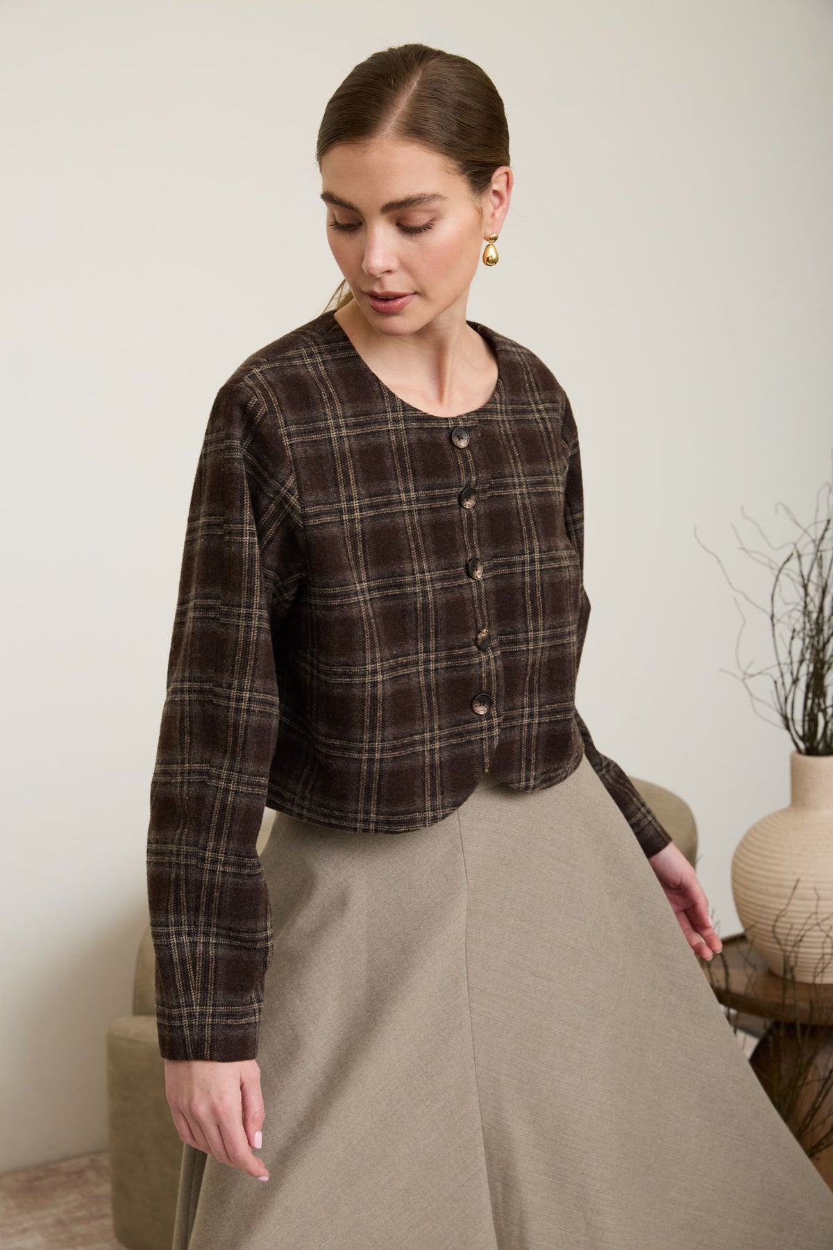A woman wearing a collarless, cropped espresso brown plaid long-sleeve jacket with tortoiseshell buttons and a beige flare skirt.