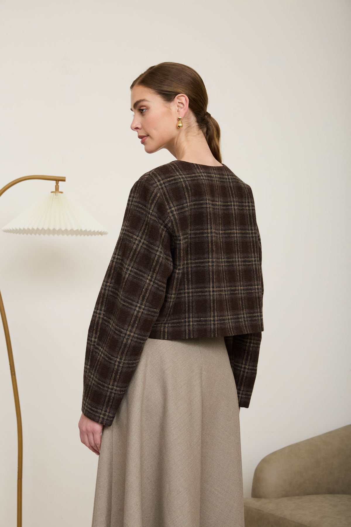 Rear view of a woman wearing a collarless, cropped espresso brown plaid long-sleeve jacket and a beige flare skirt.