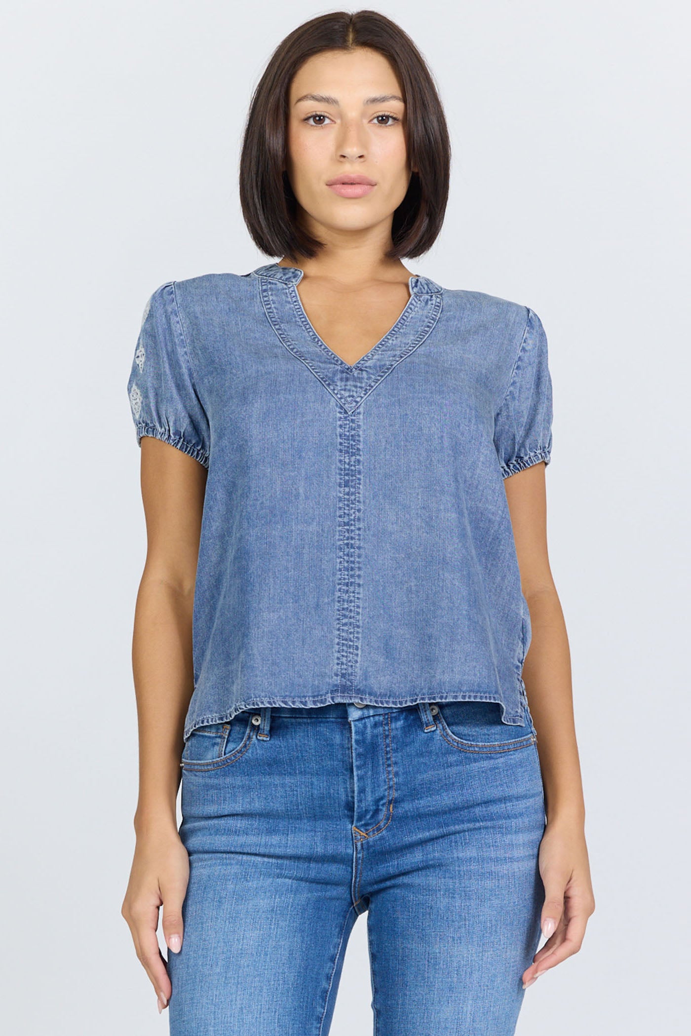 Front view of a woman wearing a light blue 100% Tencel V-neck blouse with embroidered short puff sleeves, paired with denim jeans.