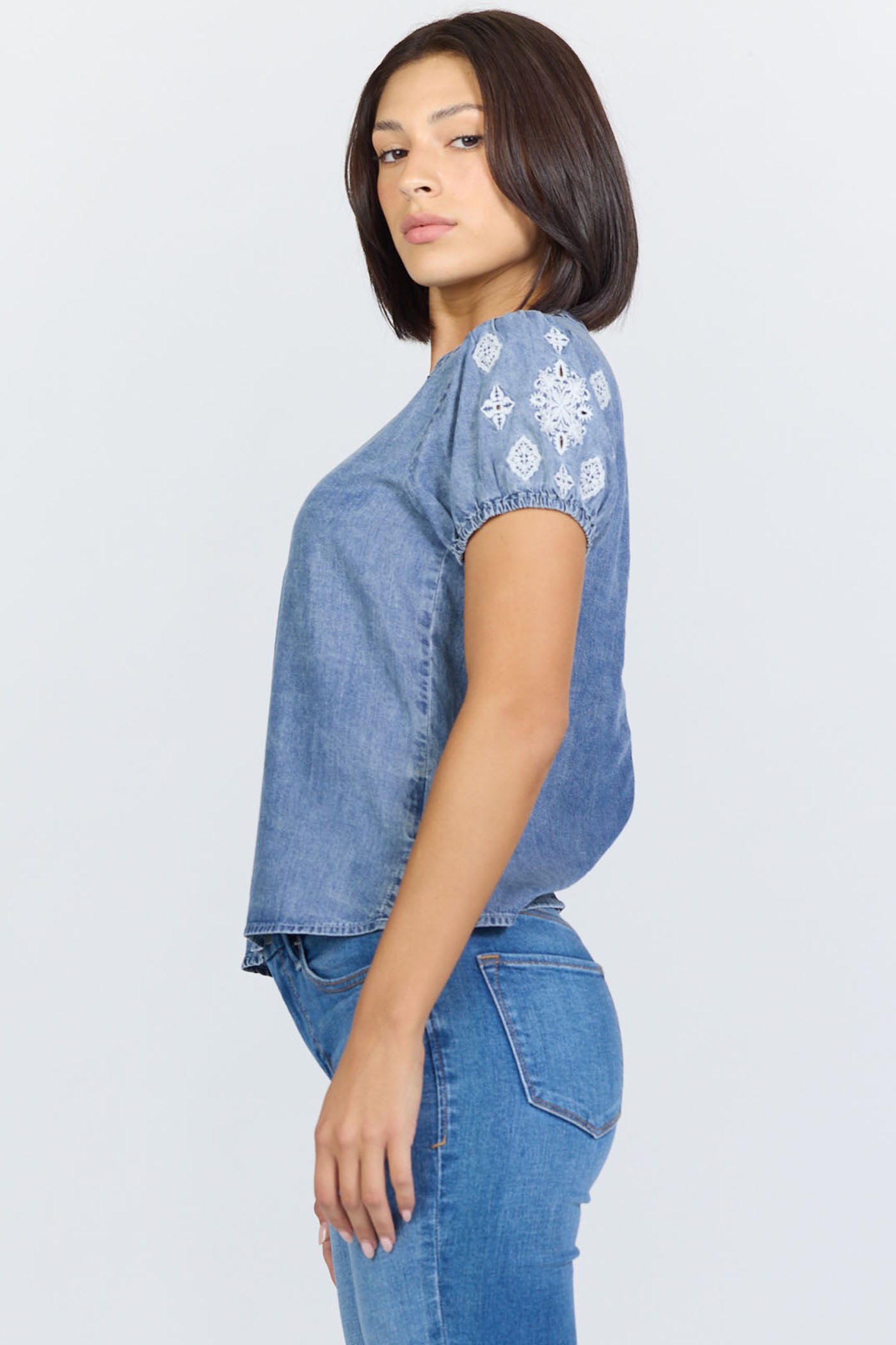 Side view of a woman wearing a light blue 100% Tencel V-neck blouse with embroidered short puff sleeves, paired with denim jeans.