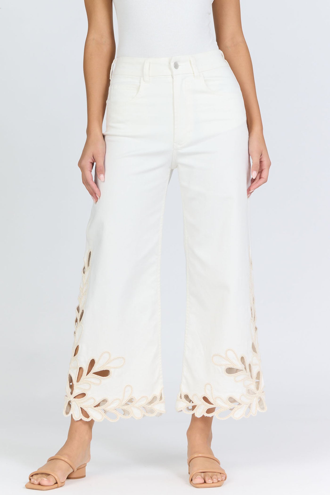 Front view of a woman wearing high-rise white wide-leg cropped jeans with an intricate floral laser-cut scalloped hemline and tan slide sandals.