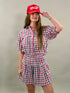 Woman smiling and looking to the side, wearing a red, navy, and white plaid short-sleeve cropped button-down shirt with a matching skort and a red baseball cap.
