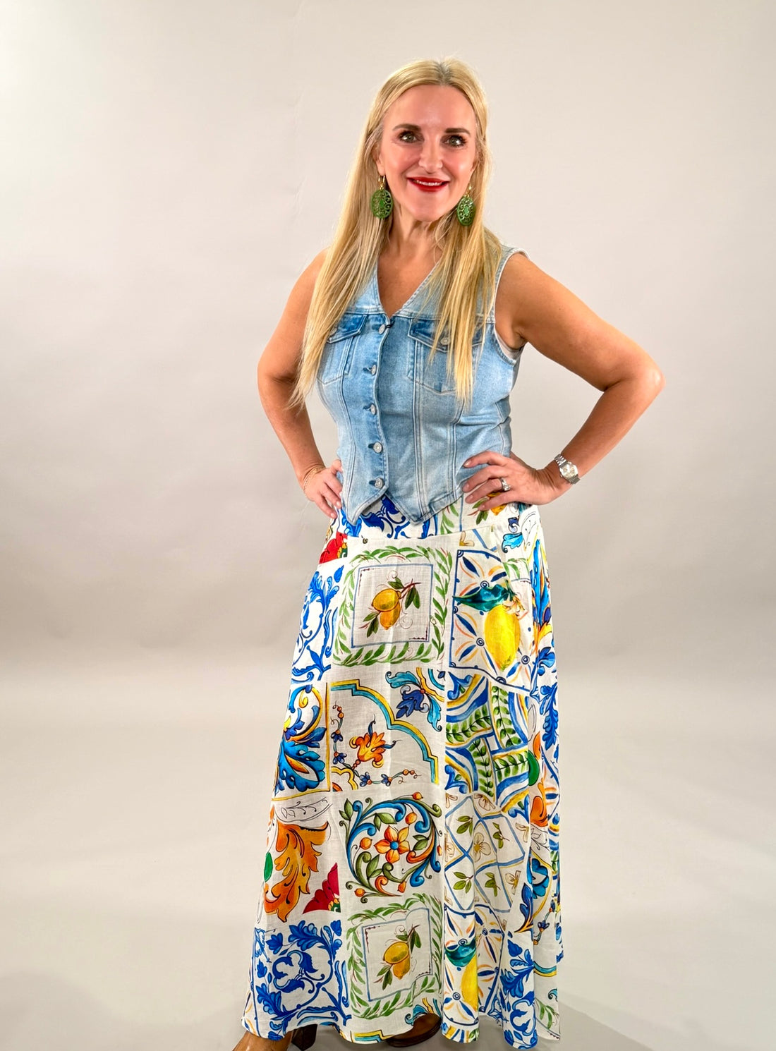 Woman standing with hands on hips wearing a light wash fitted denim vest paired with a vibrant, colorful Mediterranean-patterned maxi skirt.