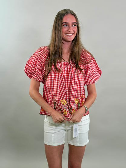 Woman looking to the side smiling while wearing a red checkered blouse with floral embroidery and white shorts against a plain background