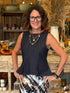 A smiling woman with short brown hair and glasses stands in a rustic home decor or pottery shop, wearing a sleeveless dark navy blue peplum top and a black, white, and brown patterned skirt.