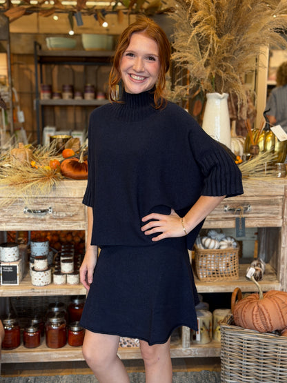 A woman modeling a dark navy blue knit mock-neck turtleneck sweater and matching mini skirt in a rustic, autumn-themed boutique setting with pumpkins and shelves in the background.