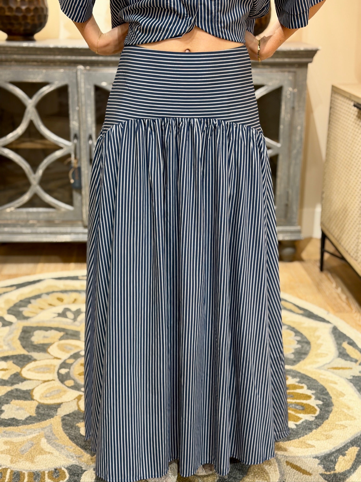 A person wearing a long navy and white vertically striped skirt with a matching striped top, standing indoors on a patterned rug in front of a cabinet.