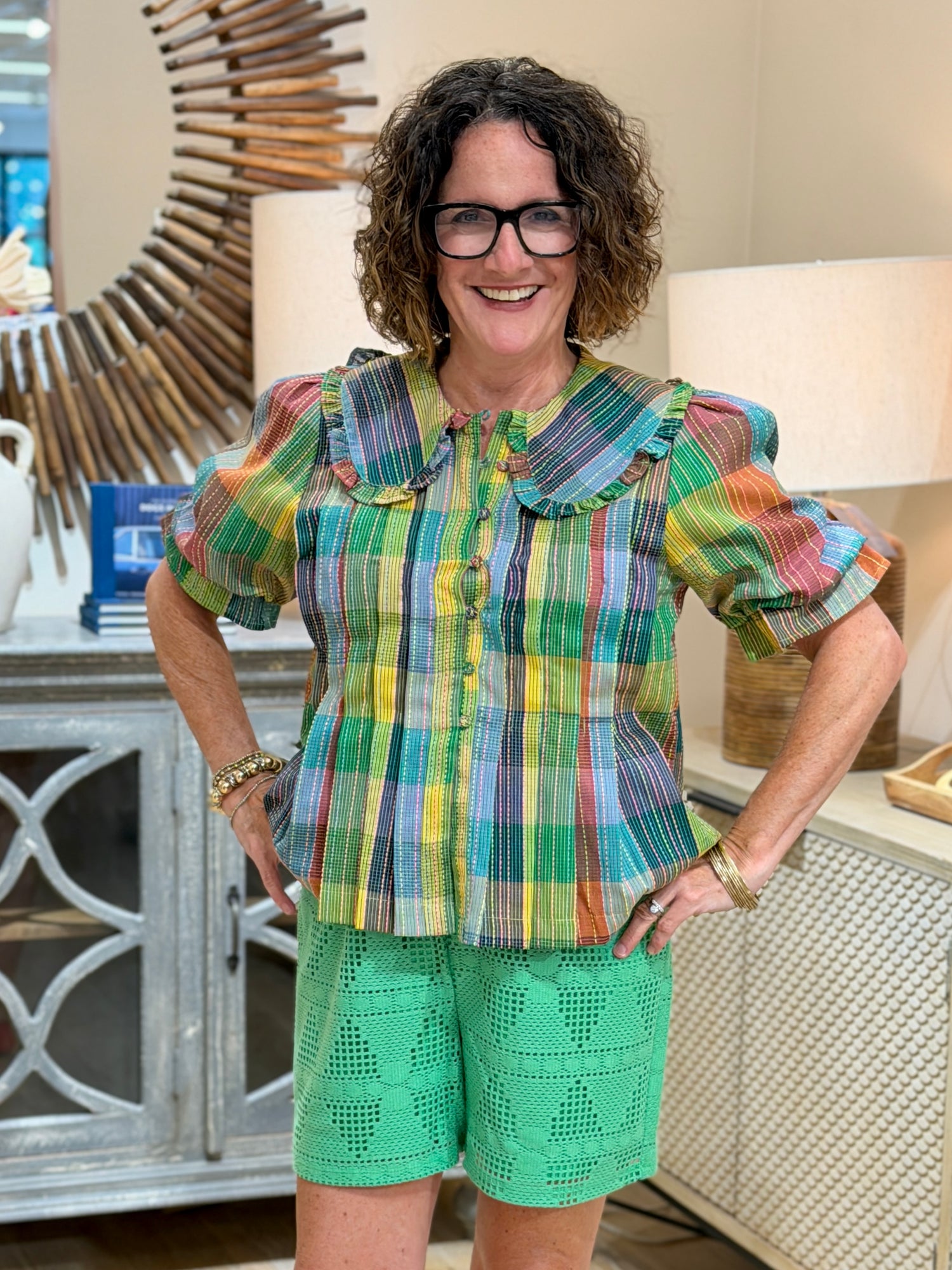 Woman with curly hair smiling while wearing a multi-colored plaid short sleeve blouse with a large ruffled collar and green eyelet shorts.