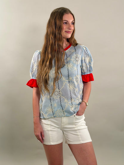 Woman looking to the side wearing a light blue floral blouse with red accents and white shorts against a plain background