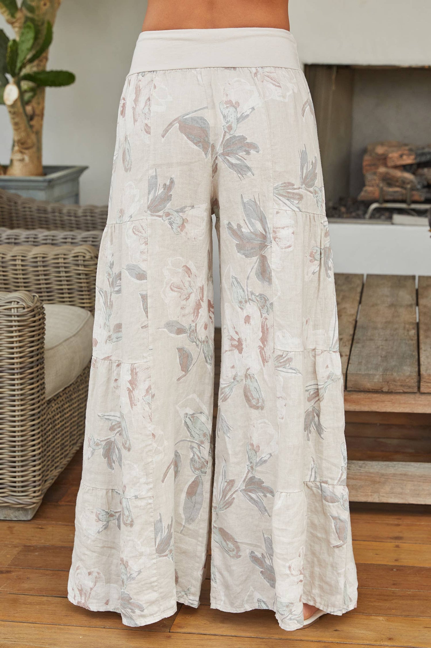 Rear view of a woman wearing cream-colored, floral-print tiered Italian linen palazzo pants with white strappy sandals.