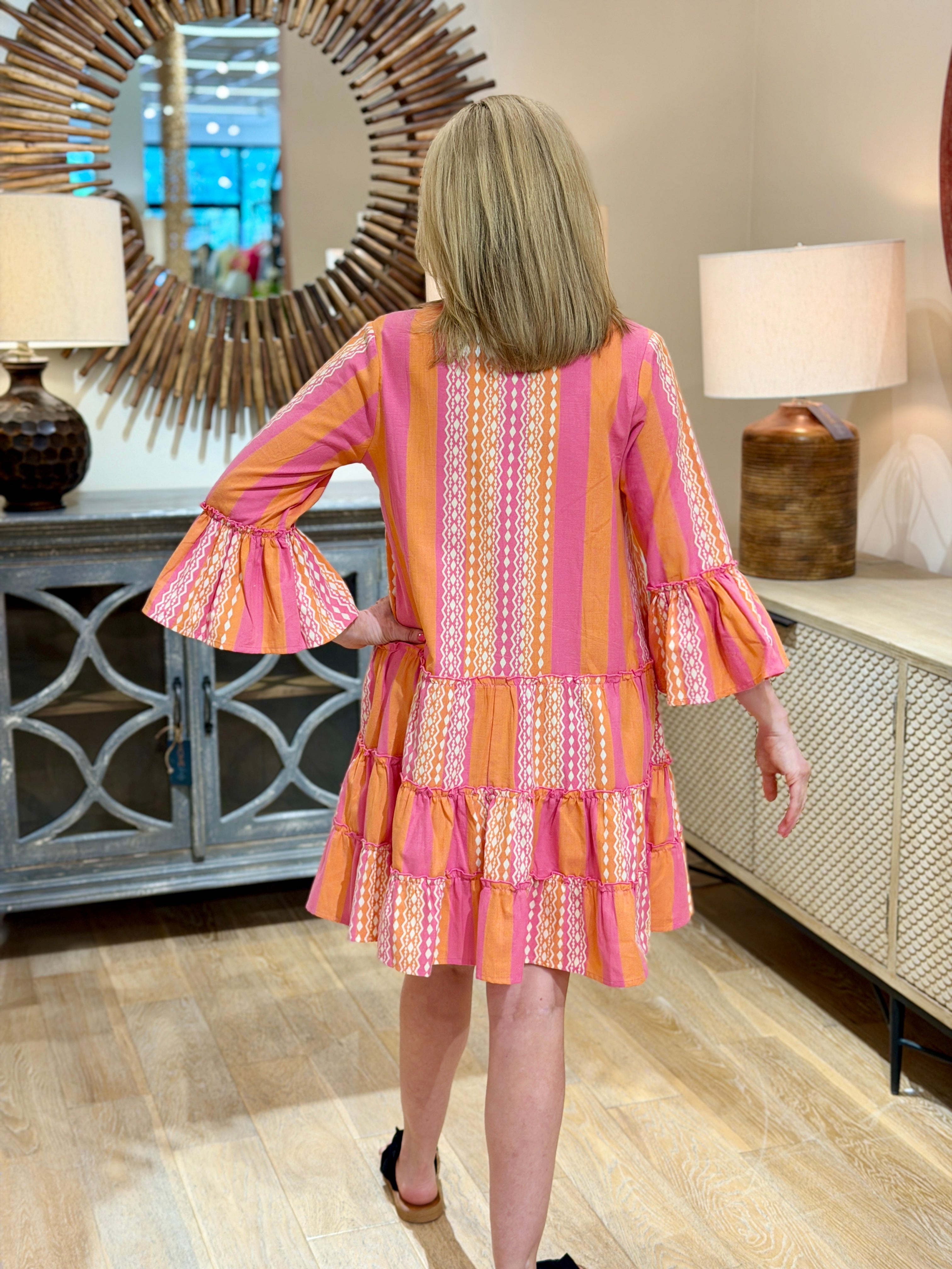 Rear view of a blonde woman wearing a pink and orange patterned 100% cotton tiered dress showing the ruffle sleeves with her hand on her hip and black slides.