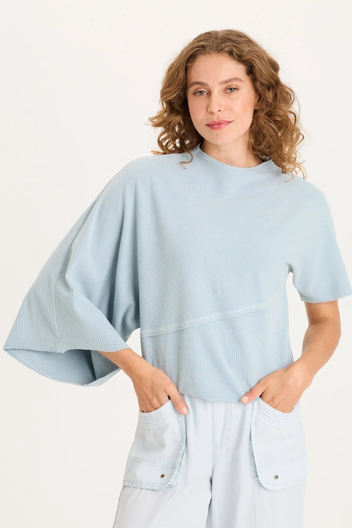 Front view of a woman with curly hair wearing a light ice-blue asymmetrical ribbed top with one flowing sleeve and one standard short sleeve.