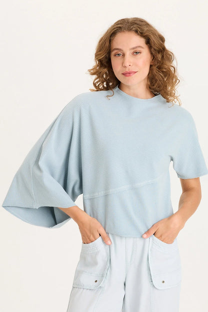 Front view of a woman with curly hair wearing a light ice-blue asymmetrical ribbed top with one flowing sleeve and one standard short sleeve.