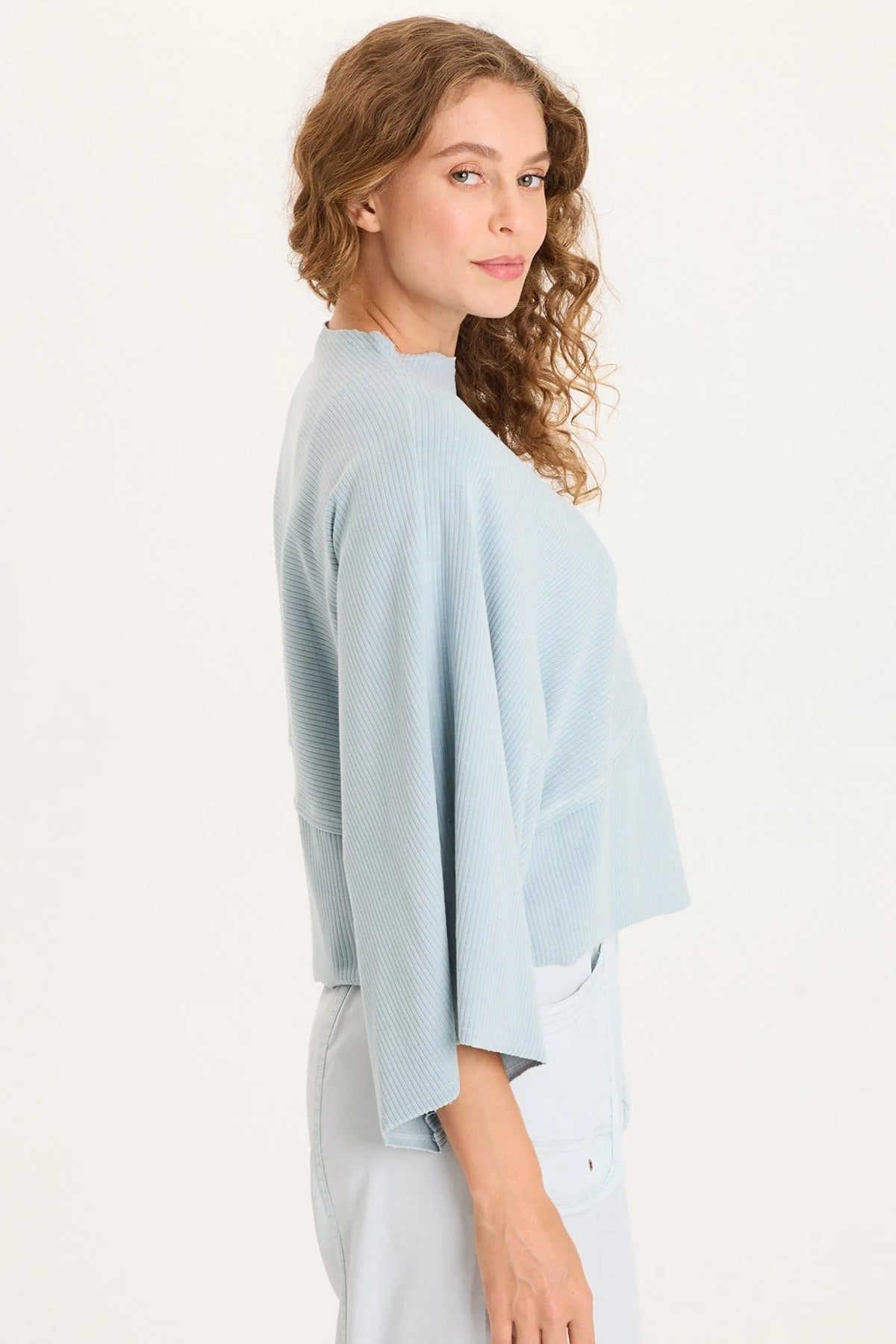 Side view of a woman with curly hair wearing a light ice-blue asymmetrical ribbed top with one flowing sleeve and one standard short sleeve.