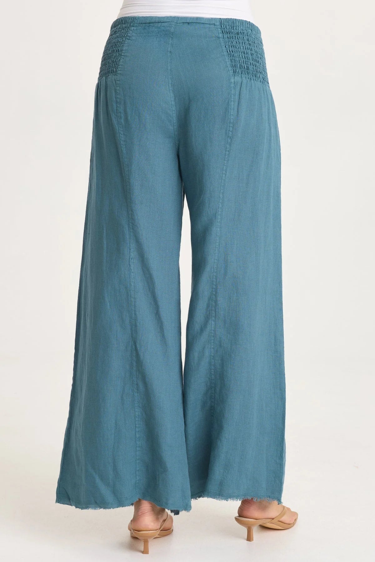 Rear view of a woman wearing flowing teal-blue wide-leg linen pants with unique smocked side waist panels and deep front pockets.