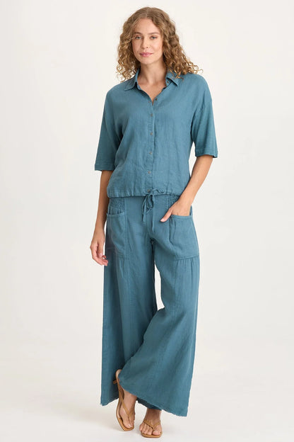 Full body view of a woman wearing flowing teal-blue wide-leg linen pants with unique smocked side waist panels and deep front pockets.